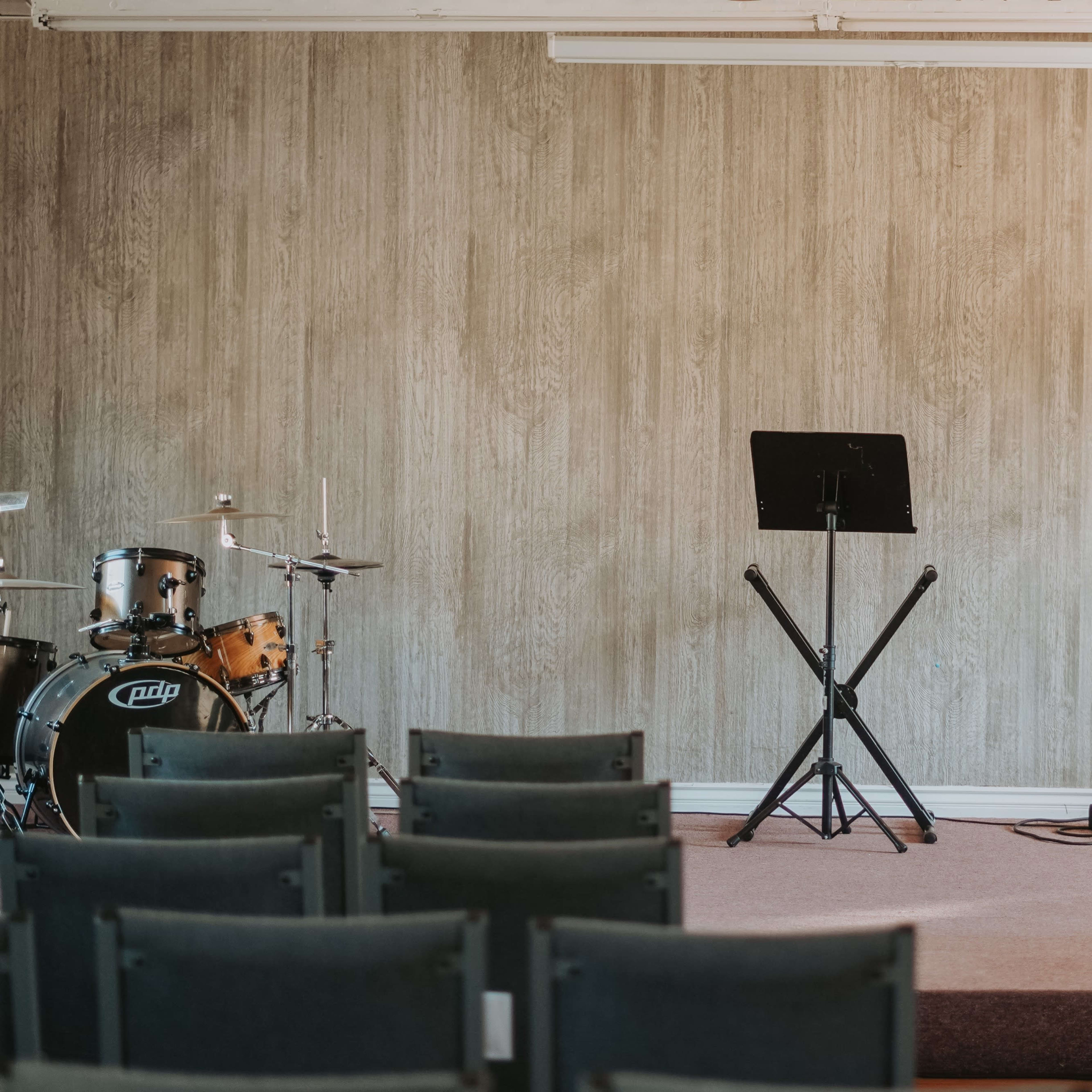 A small stage in a room features a drum set, a guitar, and audio equipment, with rows of chairs facing it.