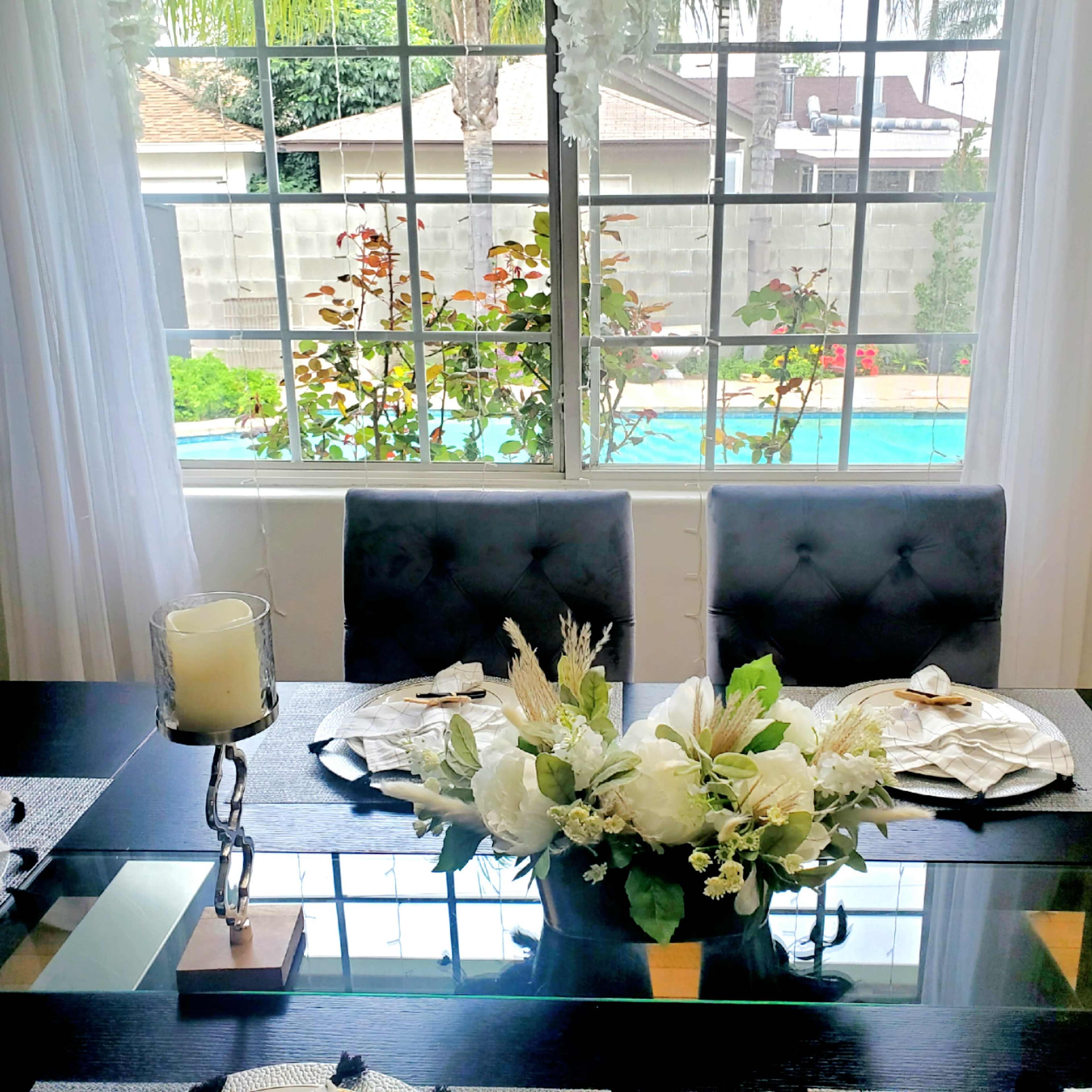 A dining table set with decorative candles and a floral centerpiece overlooks a backyard pool area through a large window.