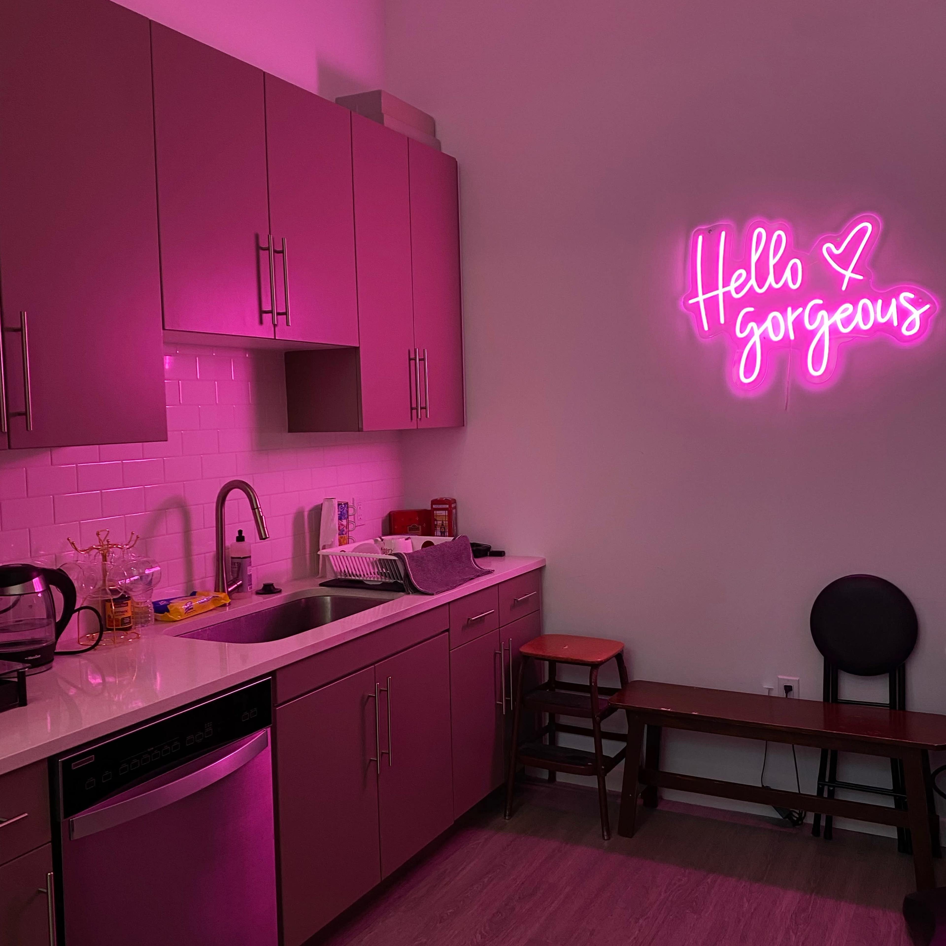 A kitchen features pink cabinets, a stainless steel dishwasher, and a neon sign that reads "Hello gorgeous" on the wall.