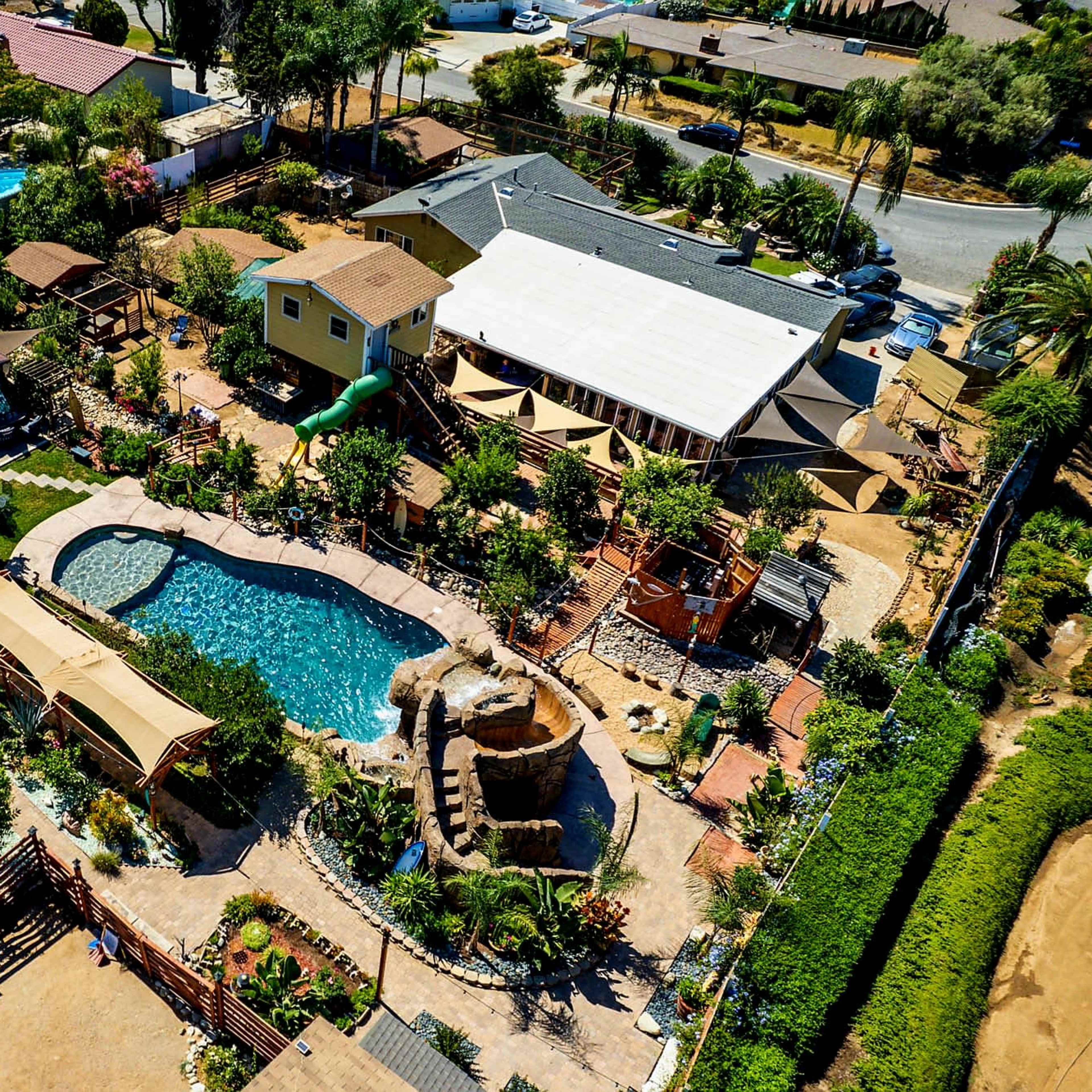 An aerial view shows a residential property with a swimming pool, landscaped gardens, and several structures, including pergolas and a water feature.