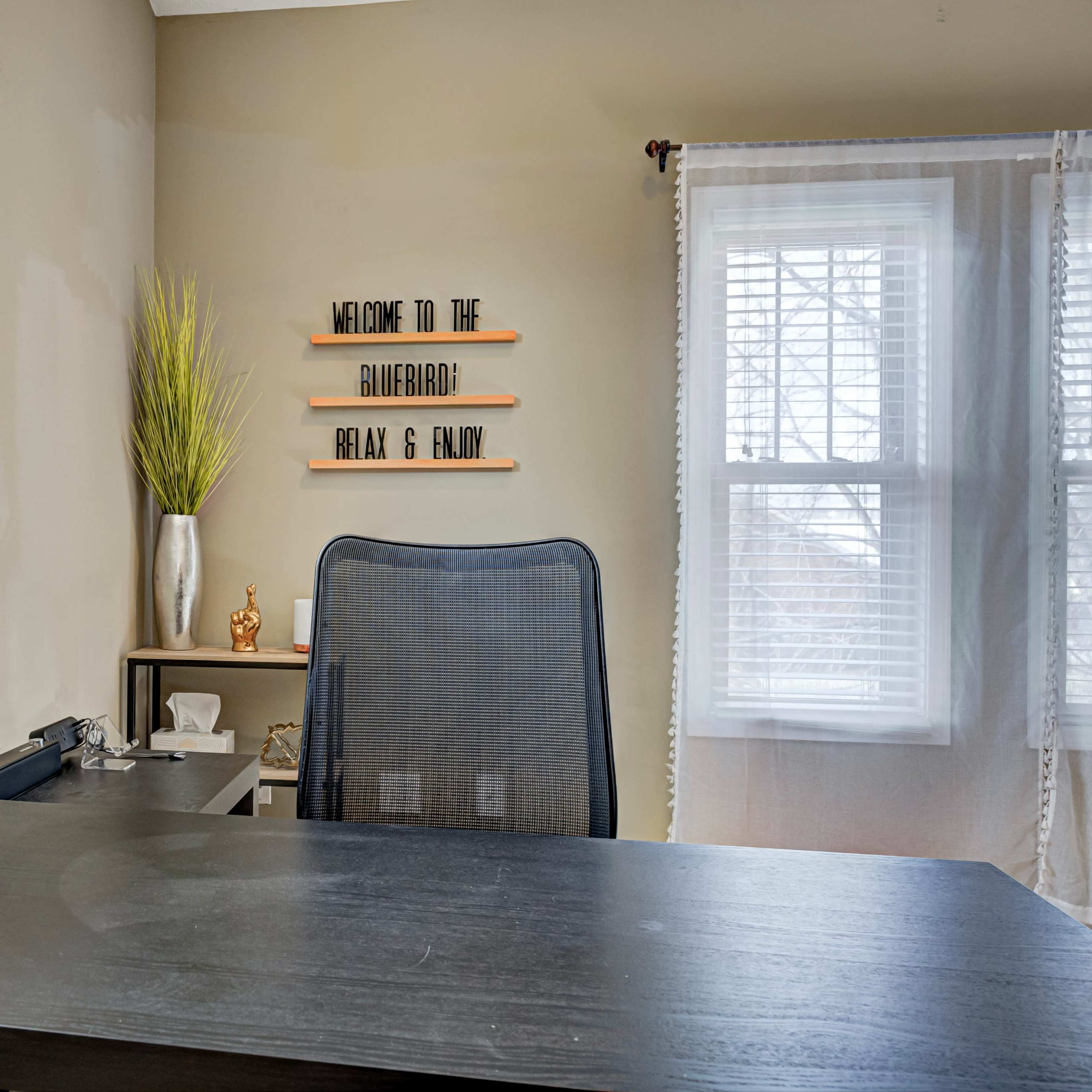 A modern office space features a black desk with a lamp, a mesh chair, and decorative elements including a plant and a welcome sign on the wall.