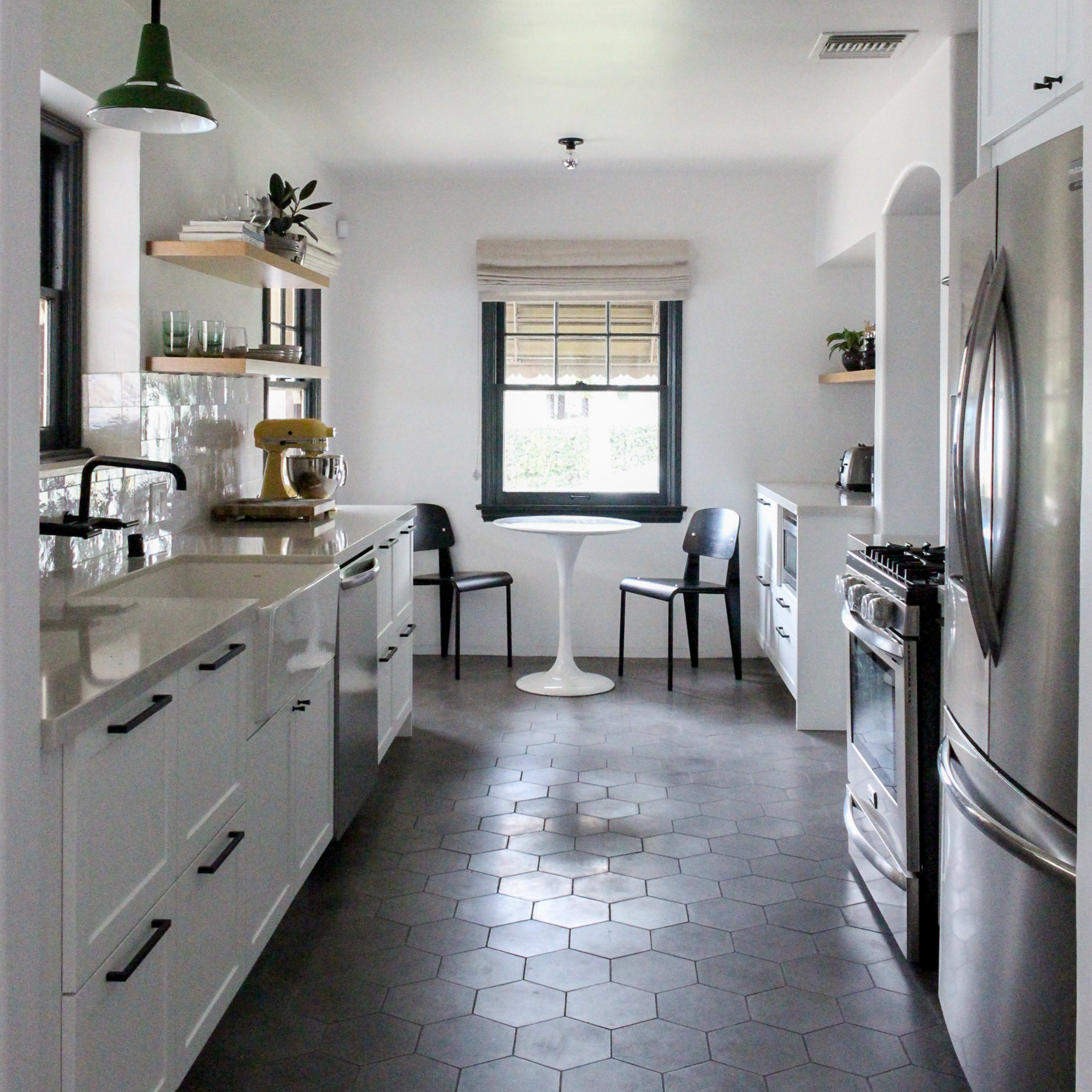 A narrow kitchen features white cabinetry, a stainless steel refrigerator, and a small table with black chairs under a bright, arching ceiling.