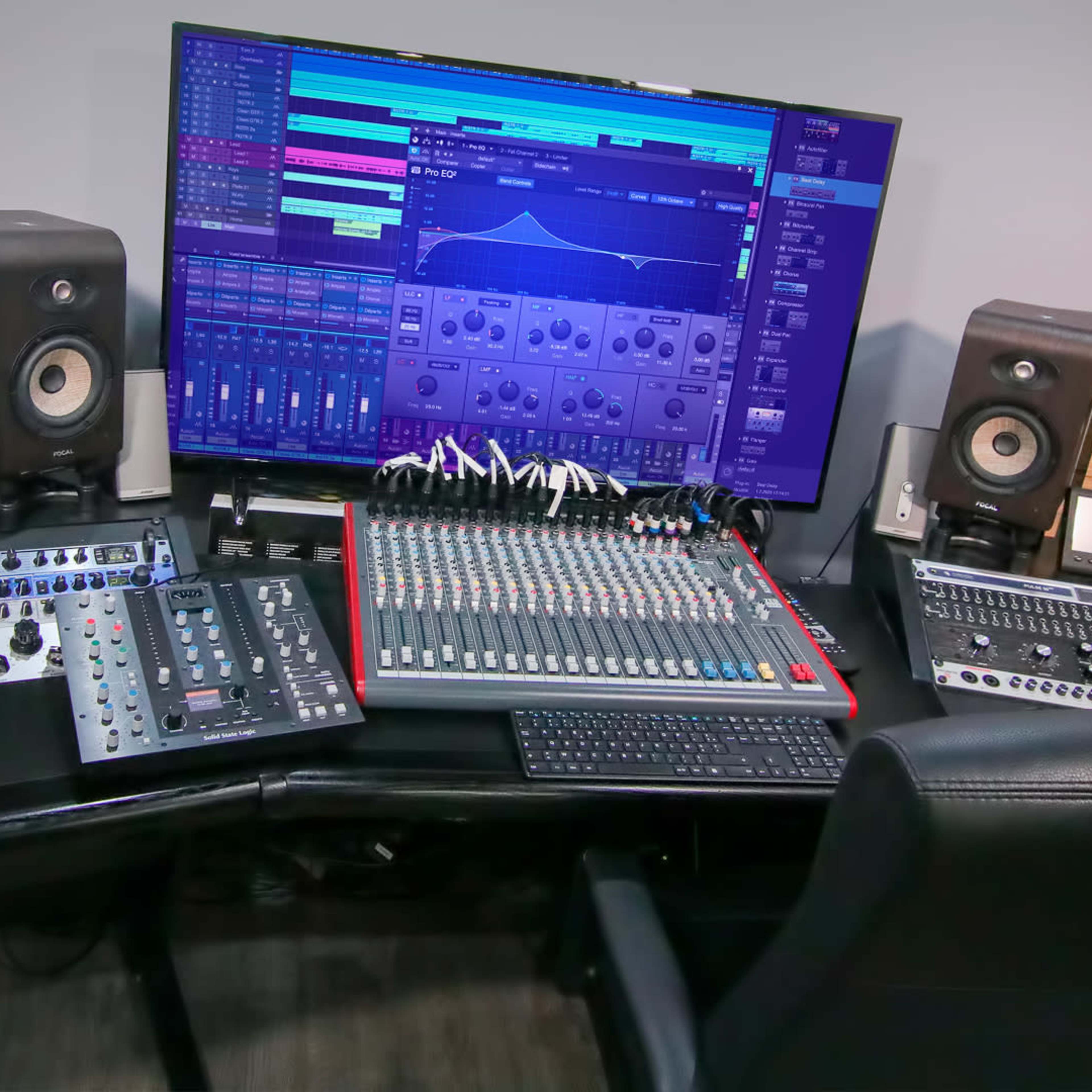 A music production setup with a large monitor displaying audio software, a mixing console, audio interfaces, and studio monitors arranged on a desk.