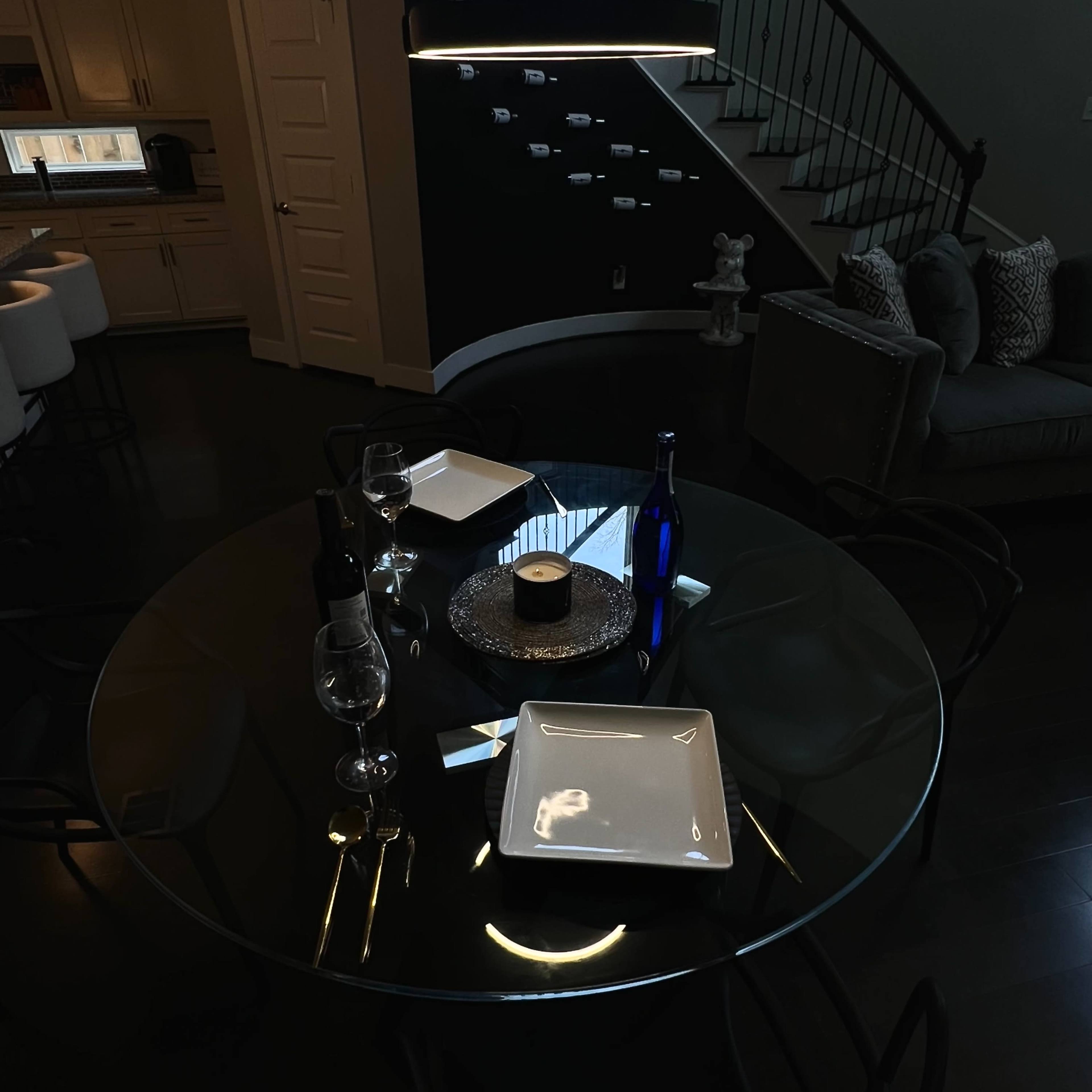 A glass dining table is set with two plates, wine glasses, and a candle, positioned next to a staircase in a dimly lit room.