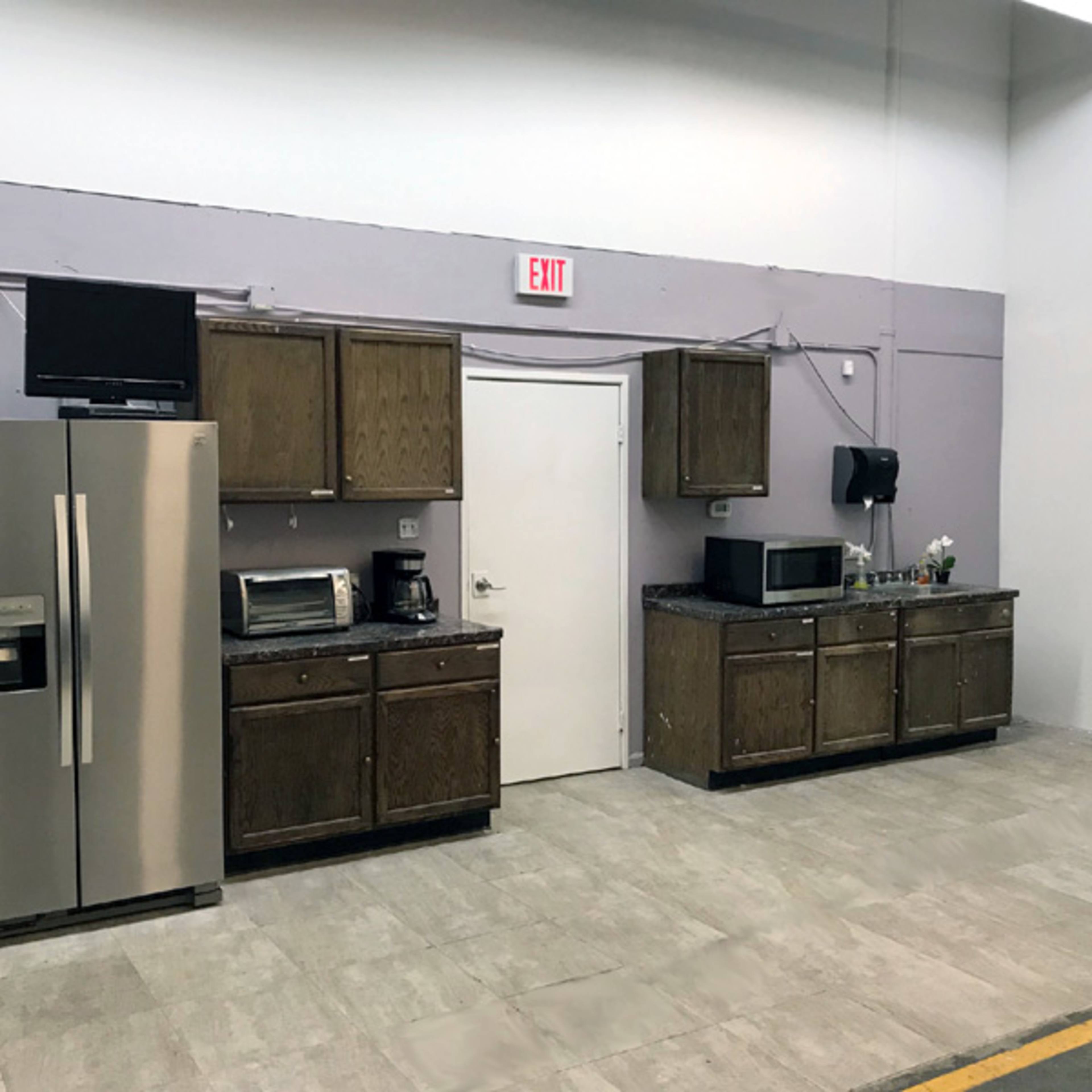 A kitchen area with a stainless steel refrigerator, microwave, coffee maker, and dark wooden cabinets against a purple wall.
