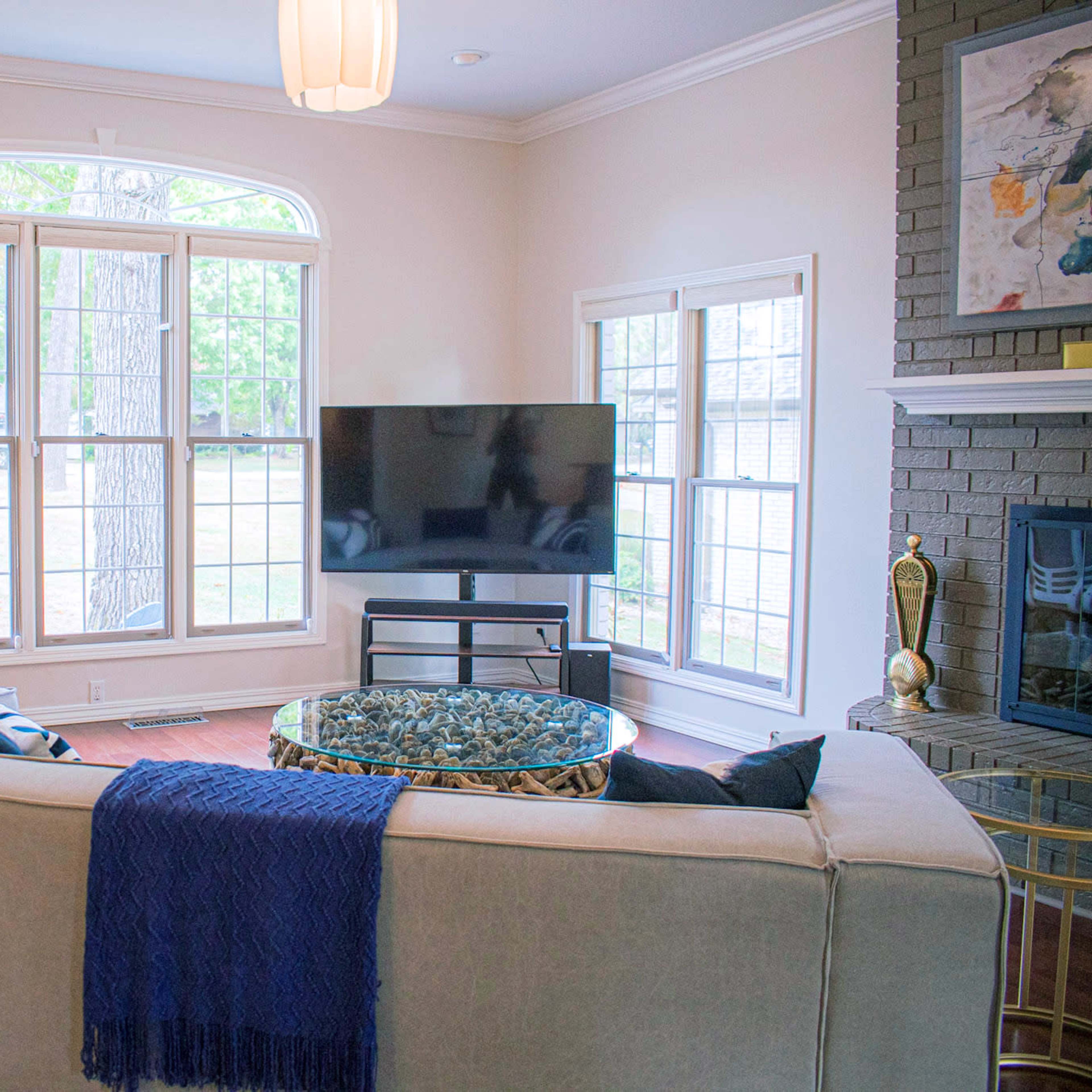 A living room features a beige sofa with a blue throw, a glass coffee table, a television on a stand, and large windows overlooking a garden.