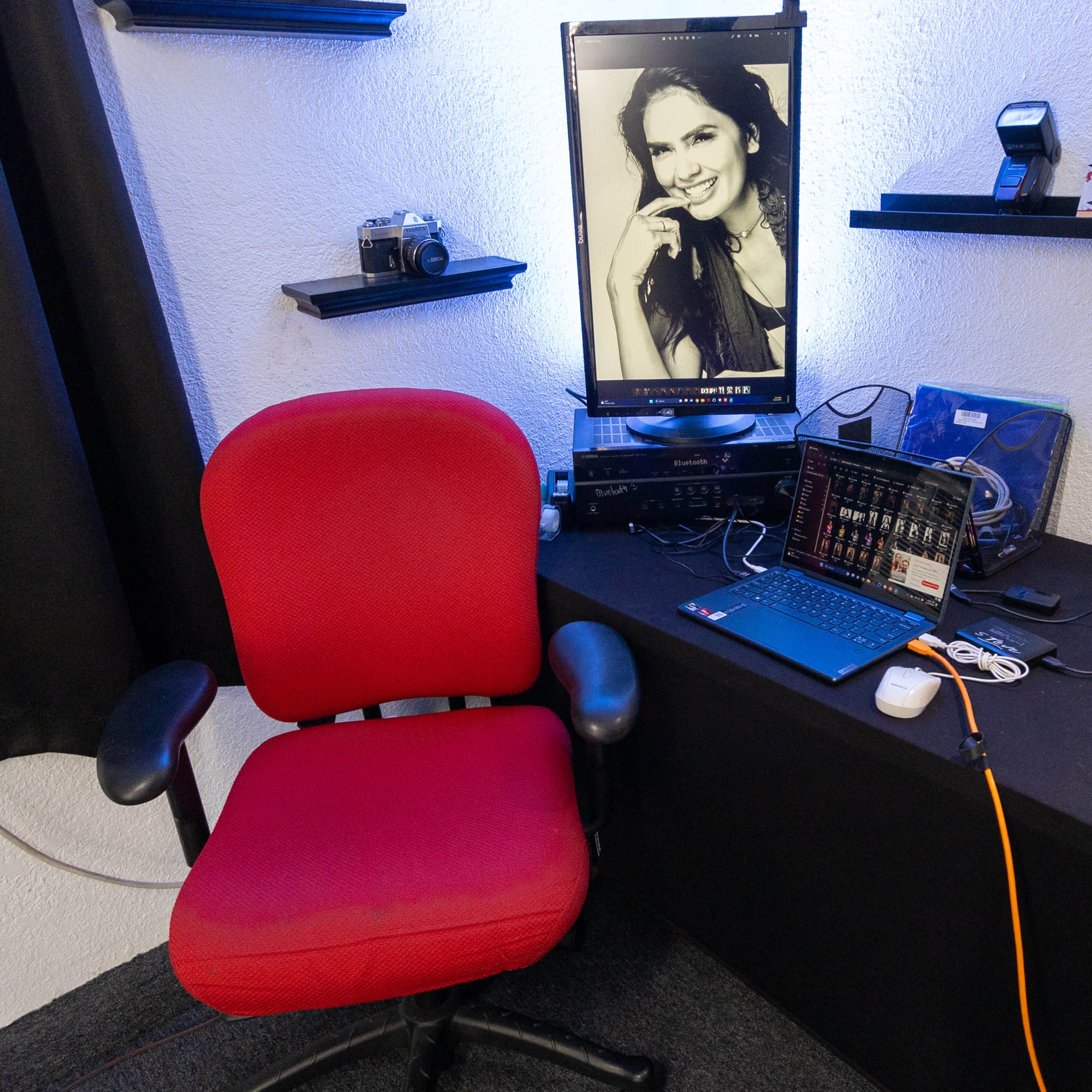 A red office chair next to a desk with a laptop, camera equipment, and a mounted monitor displaying a woman's portrait.