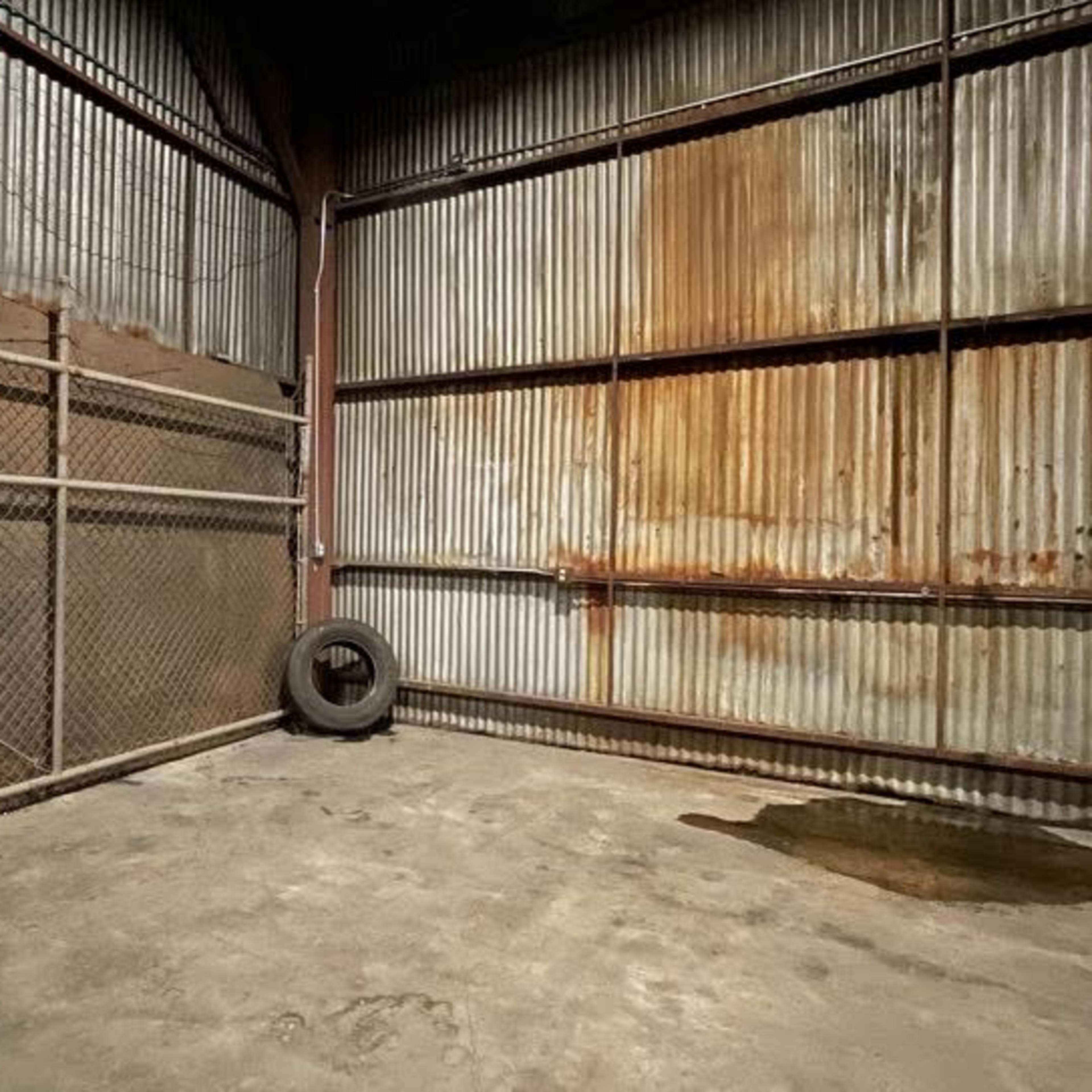 A dimly lit industrial space with corrugated metal walls, a rusted fence, and a tire in one corner.