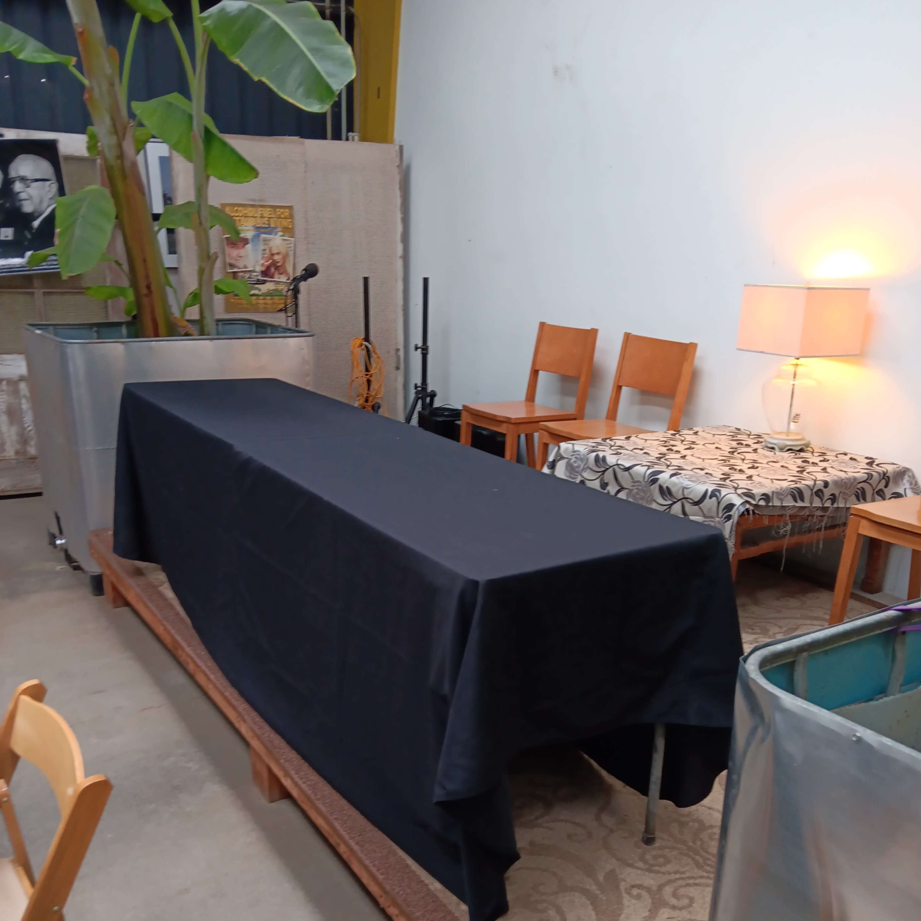 A room with a long table covered by a black cloth, surrounded by wooden chairs, a lamp, and two metal containers.