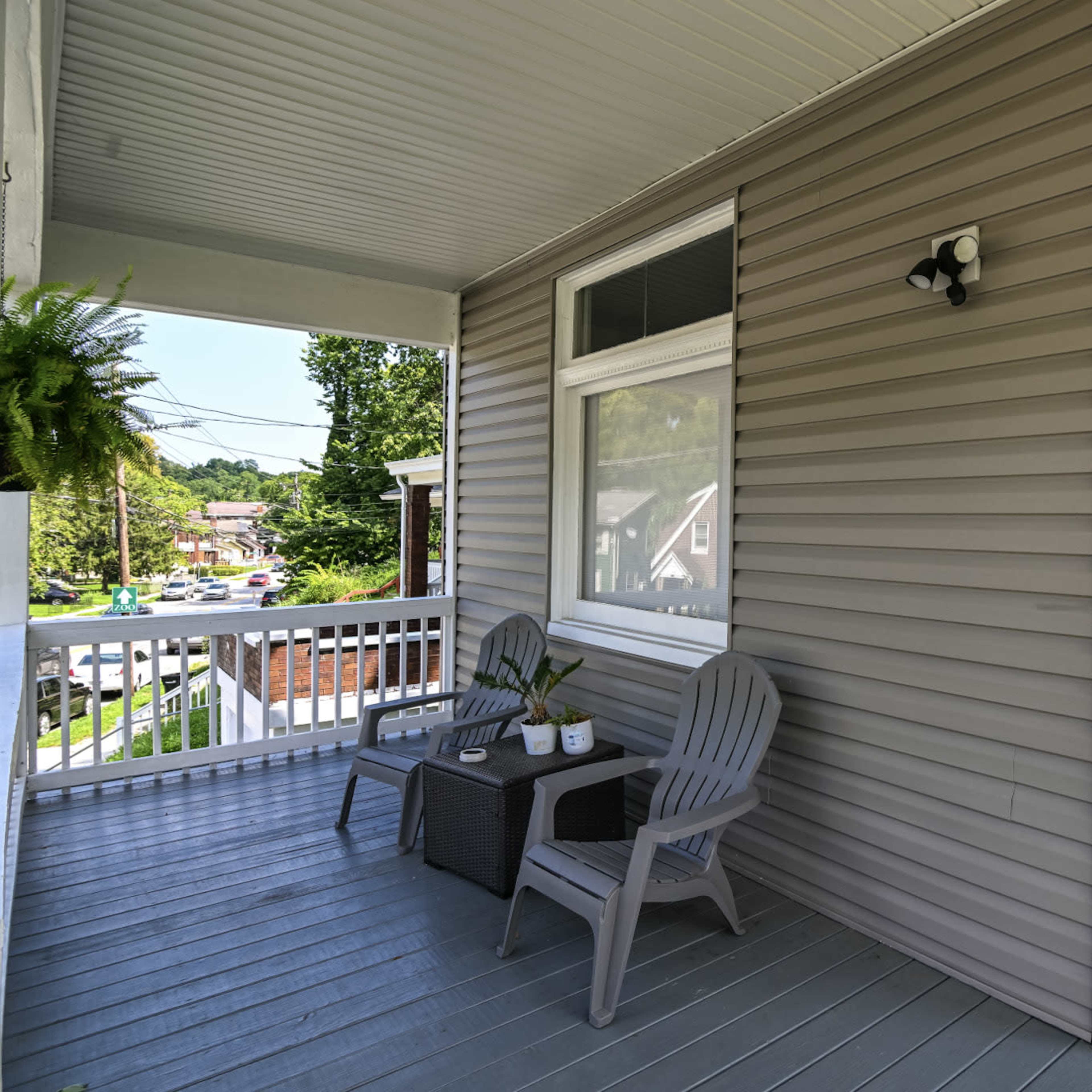 A covered porch with two gray chairs and a small table beside a window, featuring a view of a neighborhood.