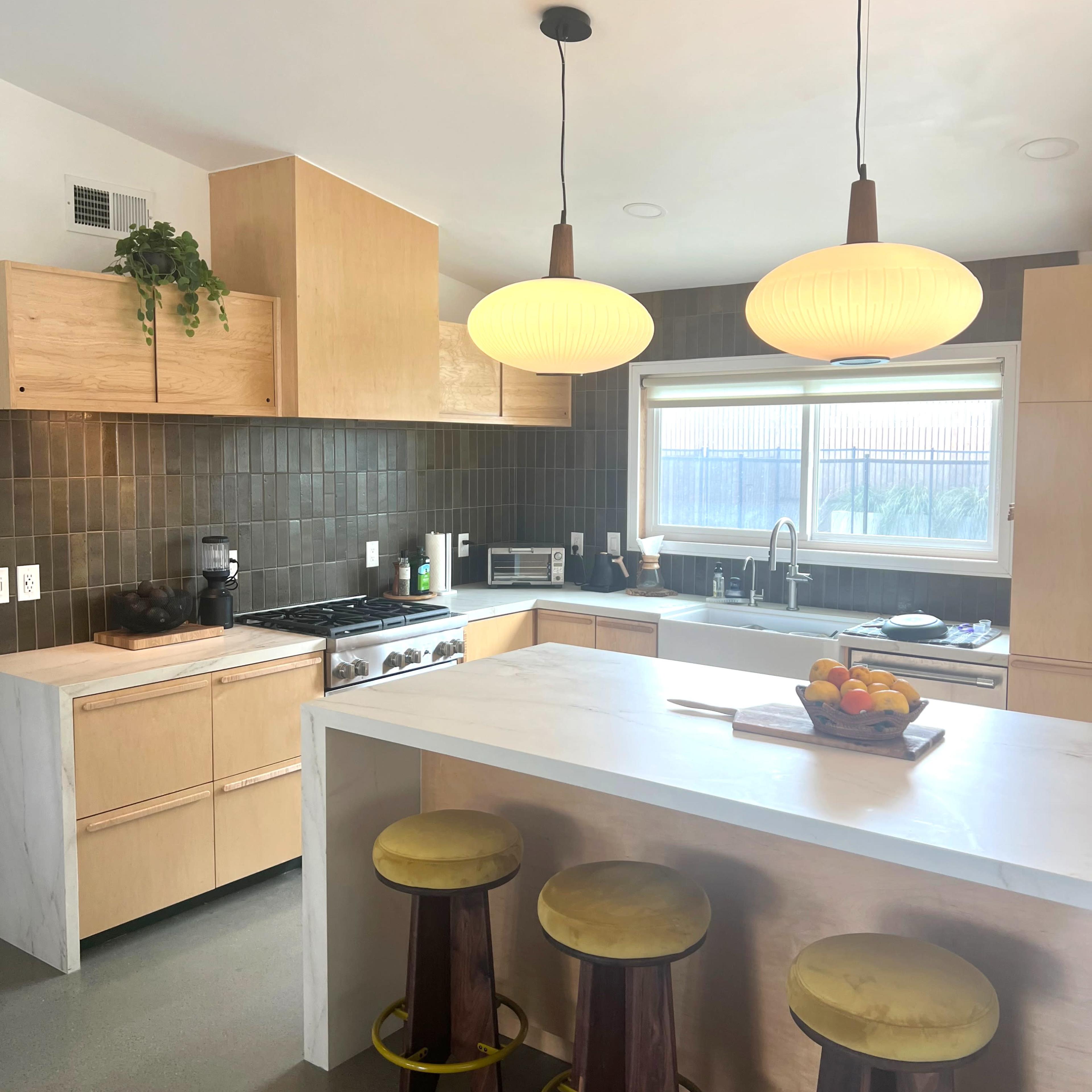 A modern kitchen with wooden cabinets, a large island with round stools, and pendant lights hanging above.