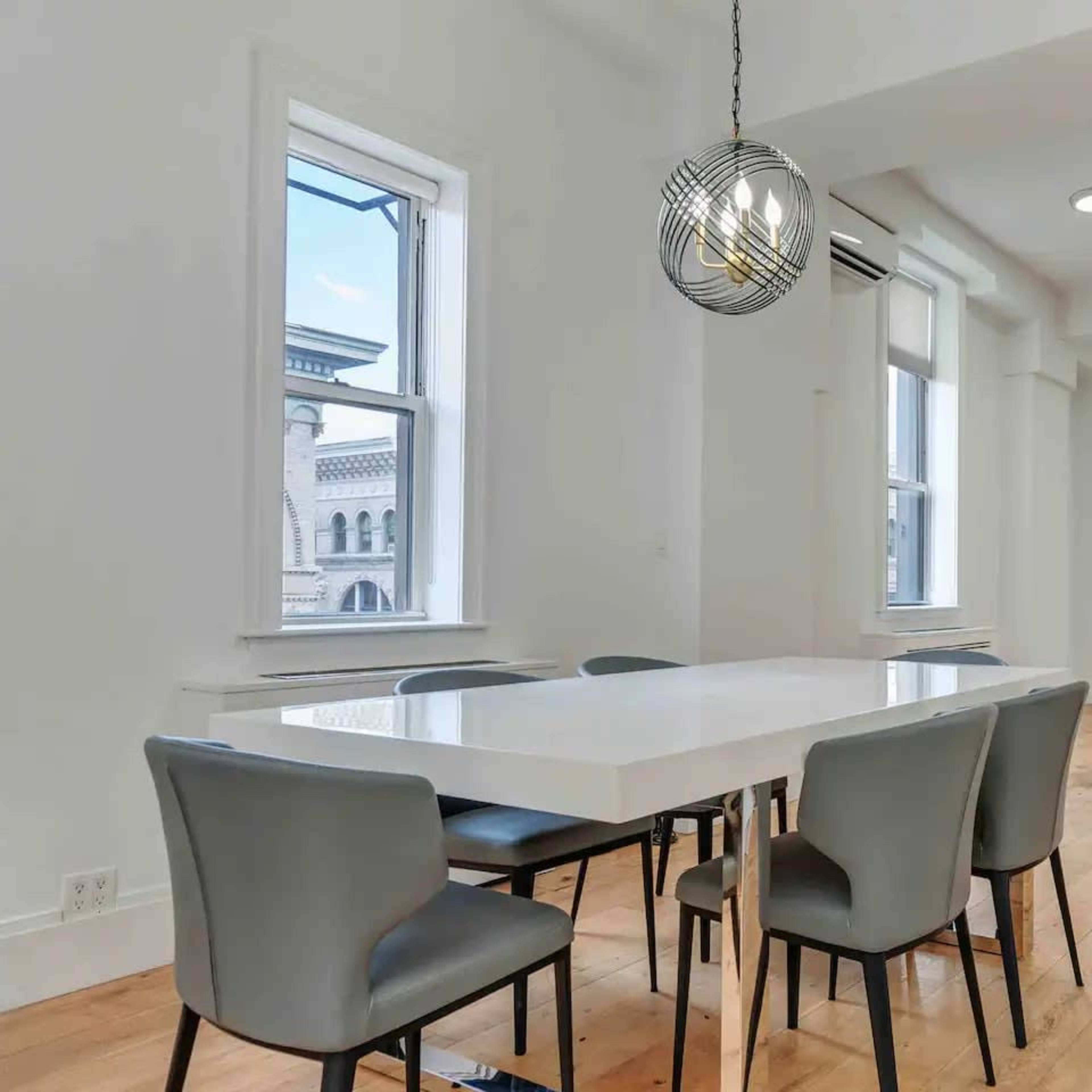 A modern dining area with a white table and gray chairs, featuring large windows and a wooden staircase in the background.