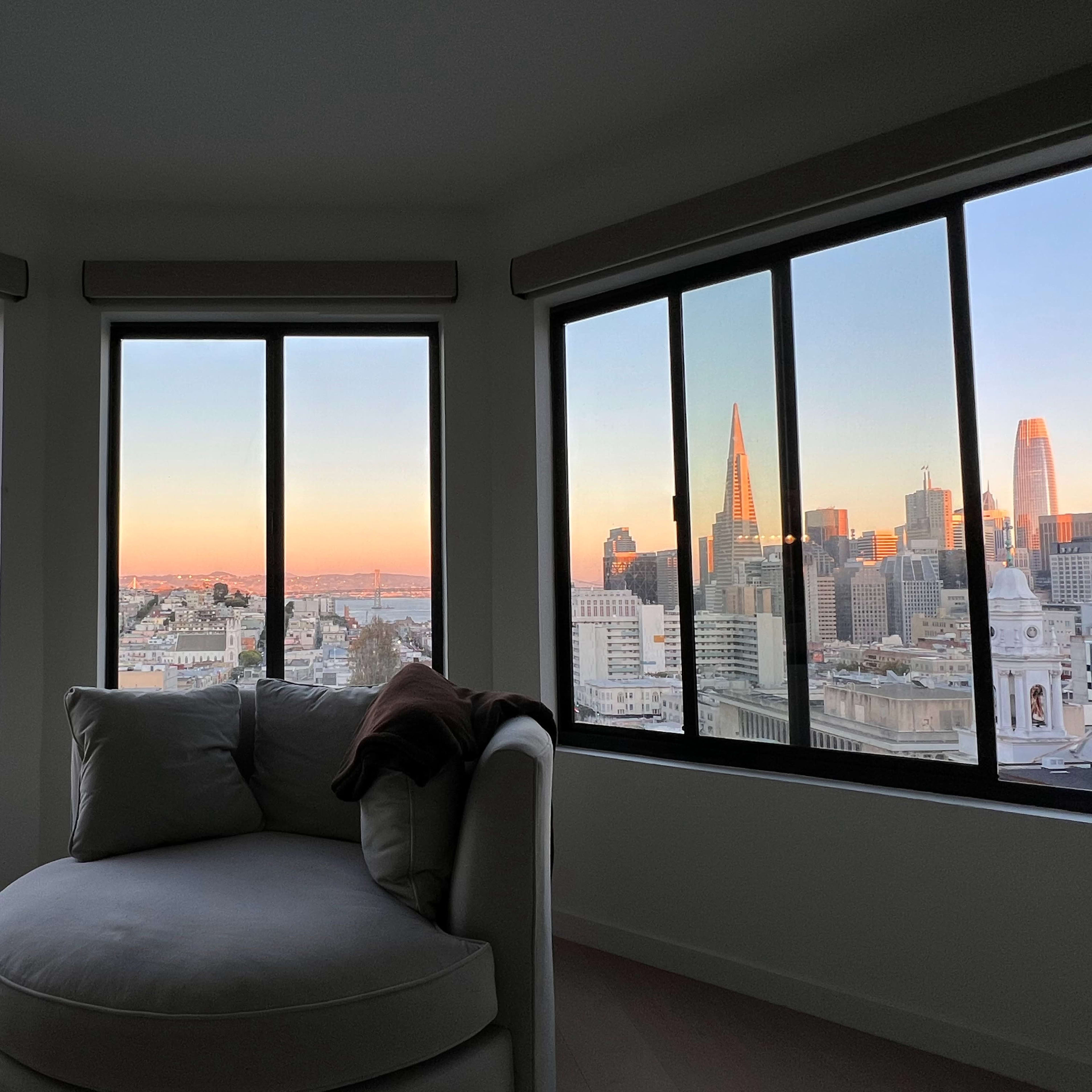 A cozy corner with a circular sofa, framed by large windows that offer a view of the San Francisco skyline during sunset.
