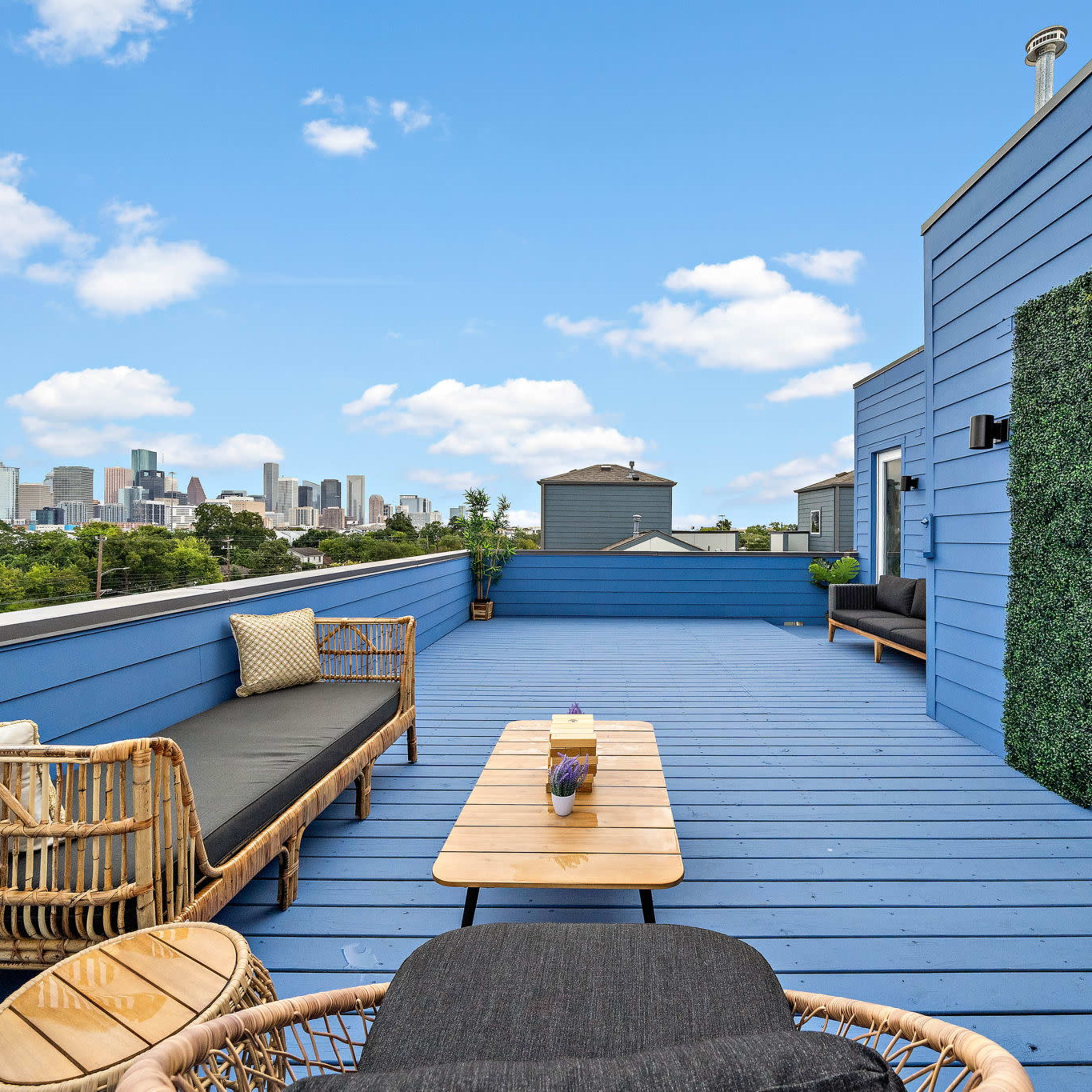 A rooftop patio features wicker furniture and a floral wall, offering a view of a city skyline under a clear blue sky.
