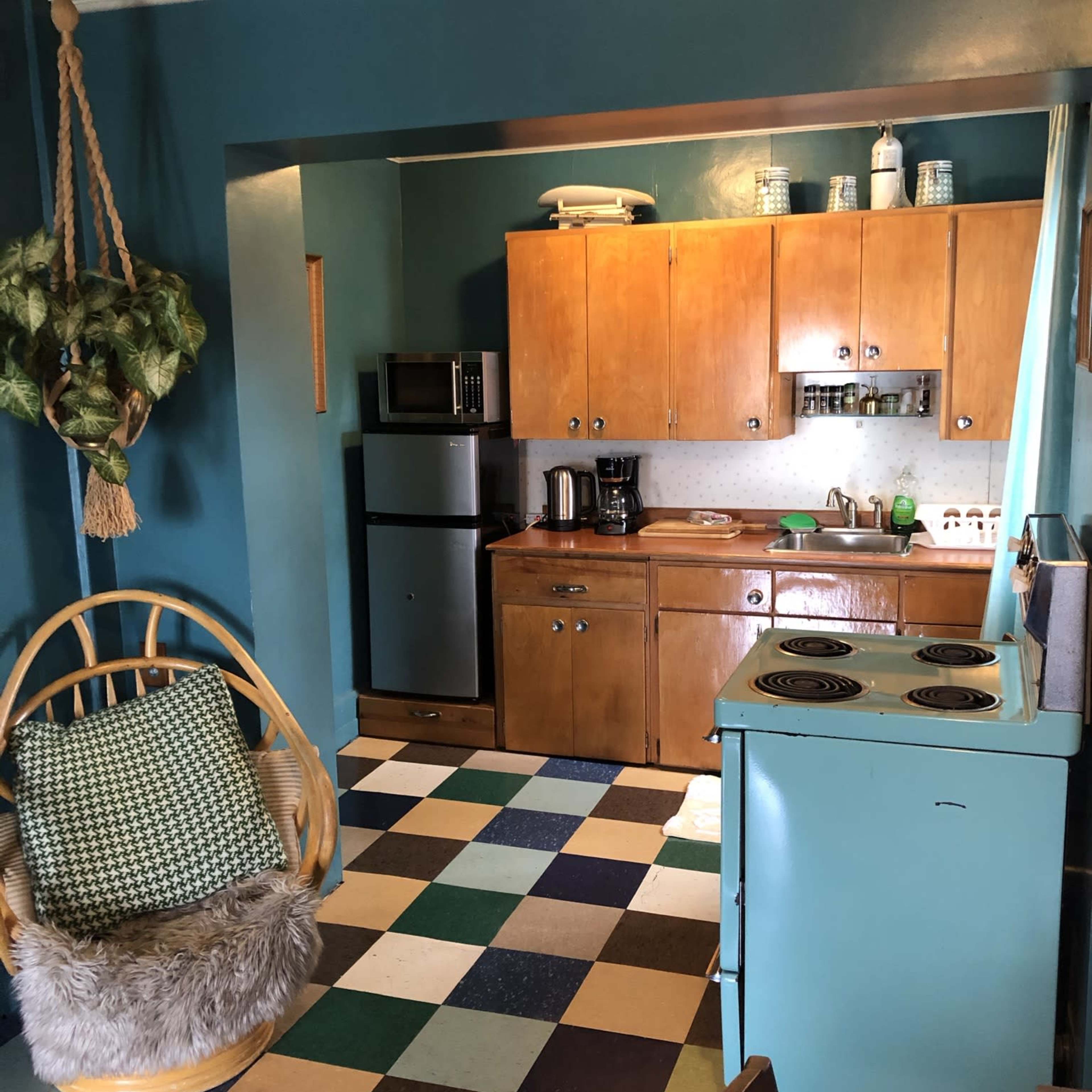 A small kitchen with teal walls, checkerboard flooring, wooden cabinets, and a seating area featuring a hanging chair.