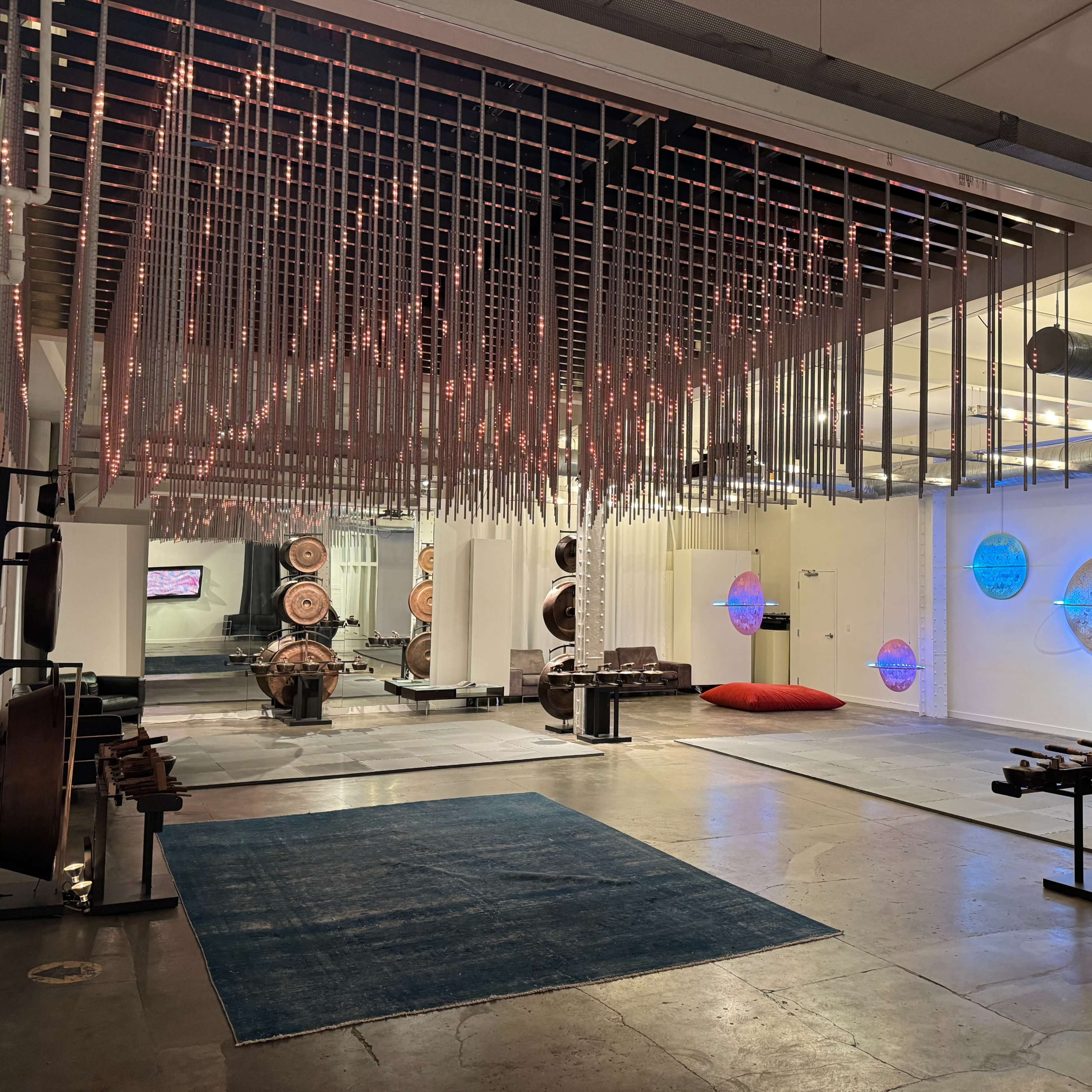 A modern, industrial space with a ceiling adorned with hanging metal rods, an open floor area featuring various musical instruments, and a large red cushion on the ground.