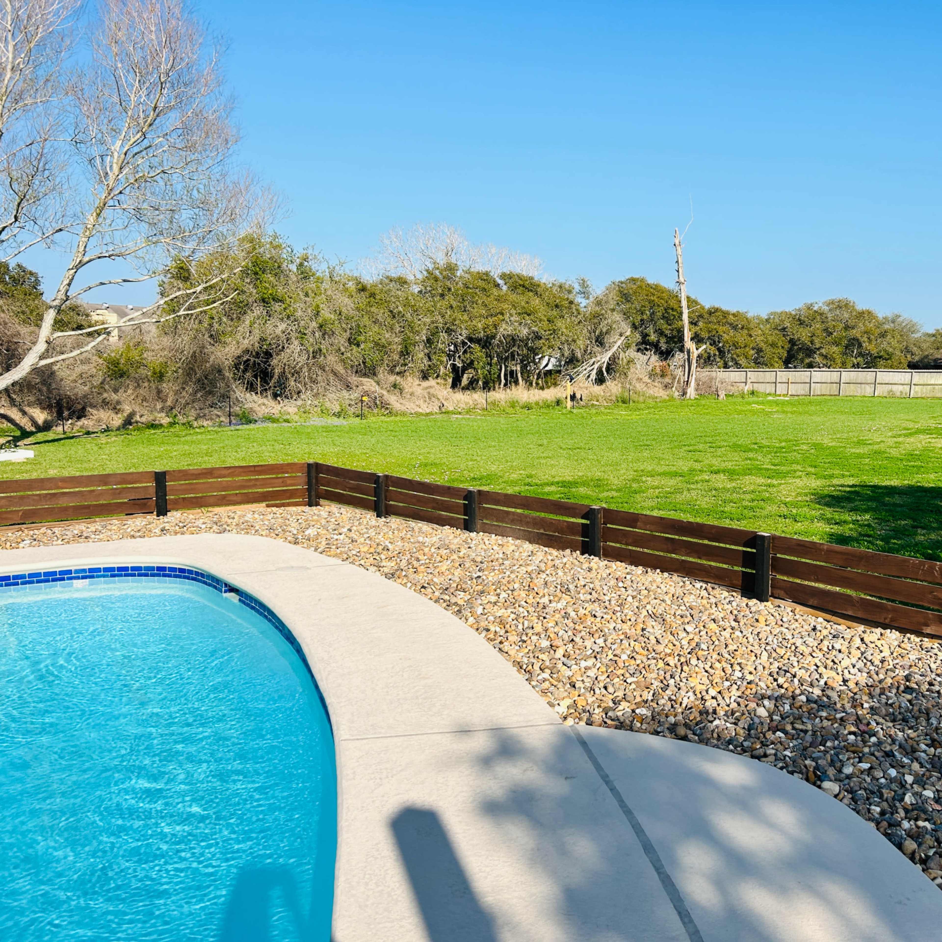 A swimming pool with a stone border is adjacent to a manicured grassy area surrounded by trees and a wooden fence.