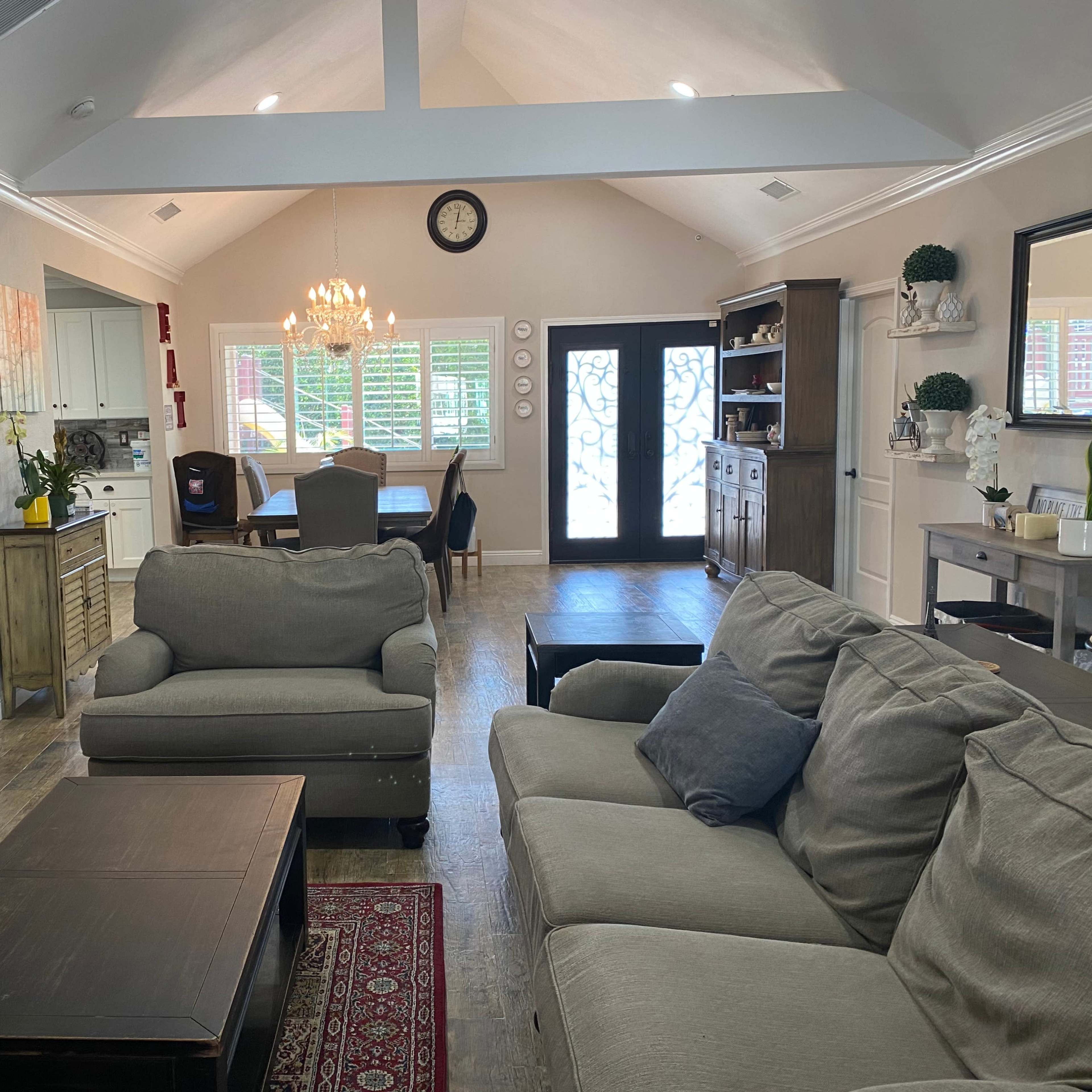 A living room features a gray sofa and coffee table, with an adjacent dining area visible through a set of double doors, and light wood flooring throughout.