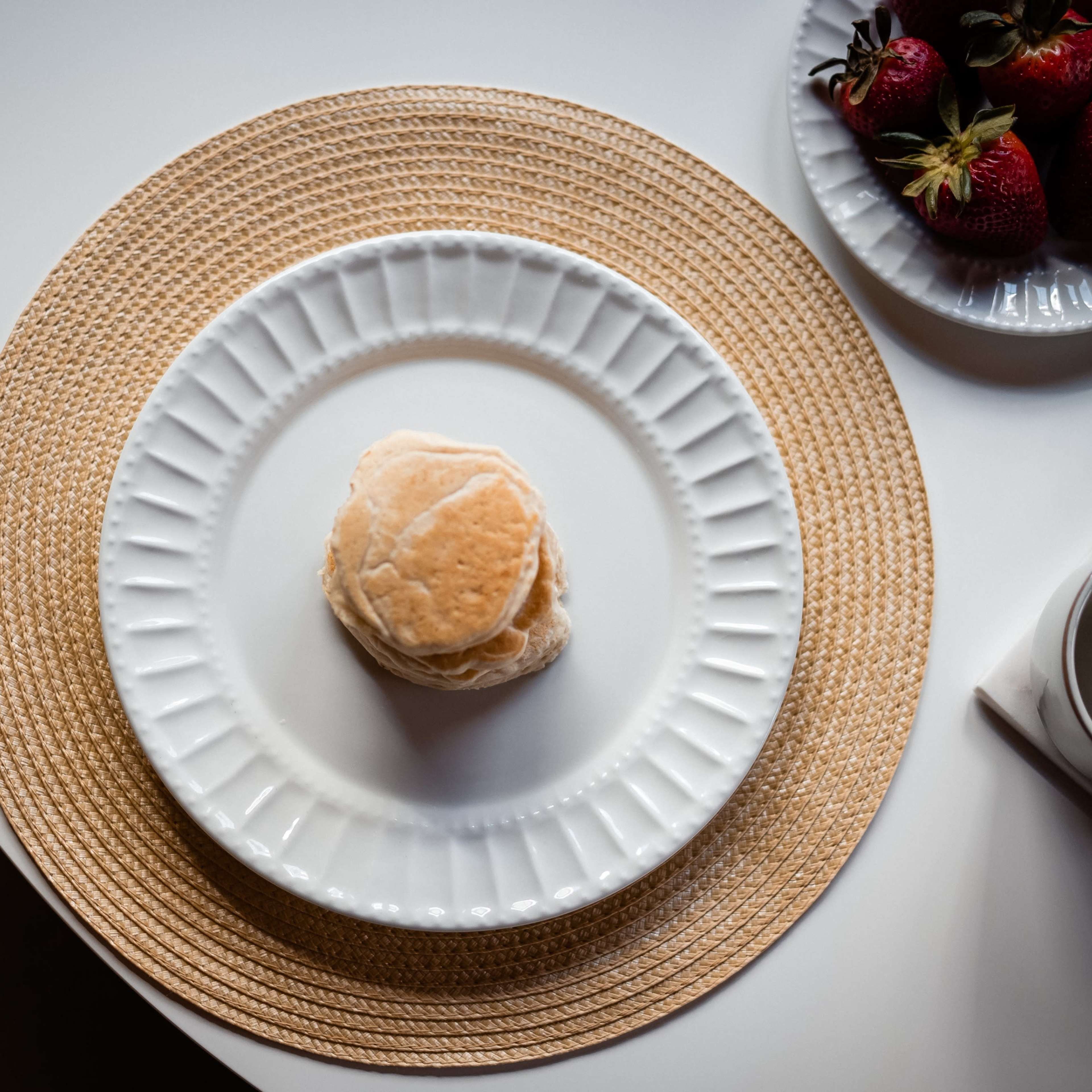 A stack of three biscuits sits on a white plate placed on a woven placemat beside a bowl of strawberries and a cup.