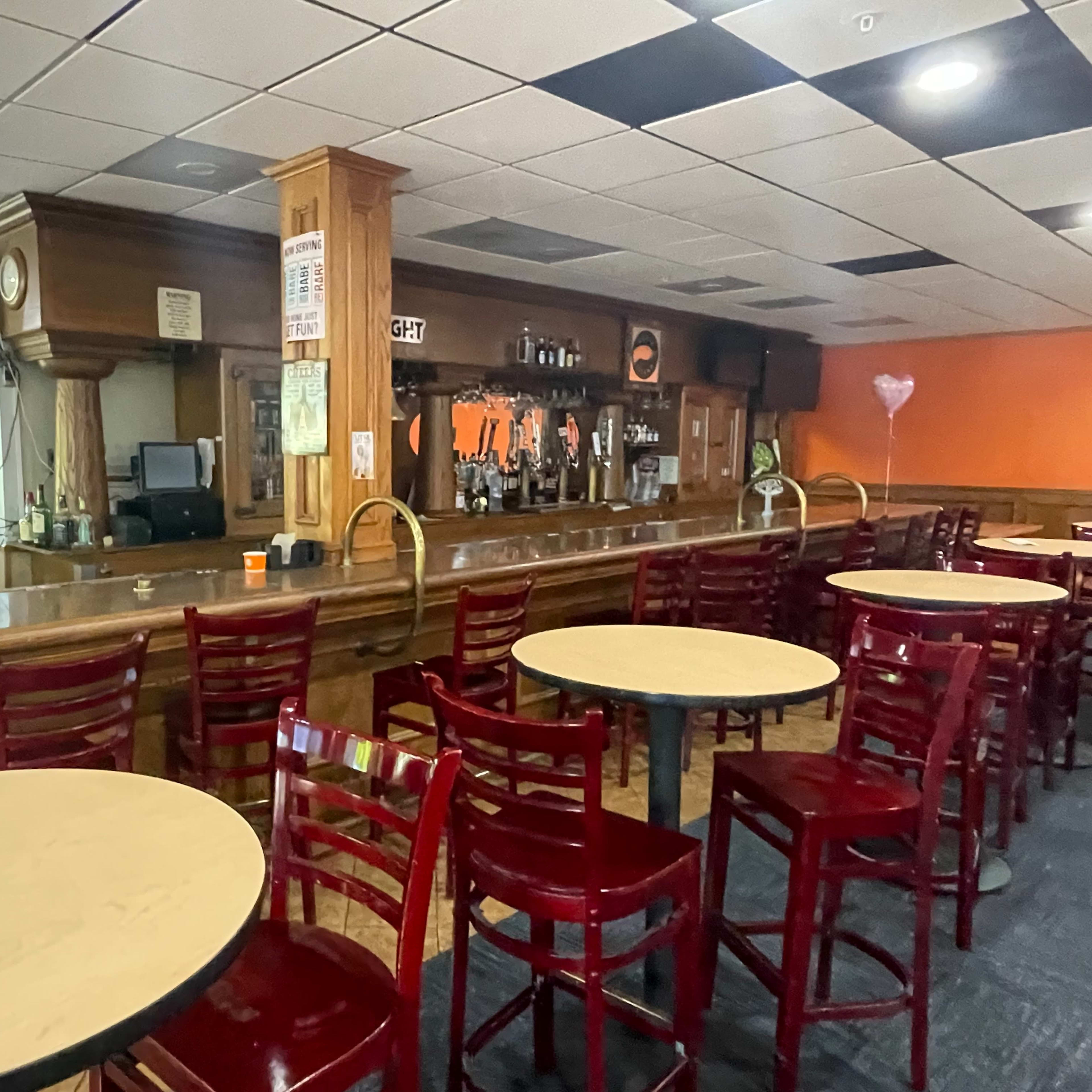 The interior of a bar features a wooden counter with empty round tables and red chairs, while shelves of bottles line the back wall.