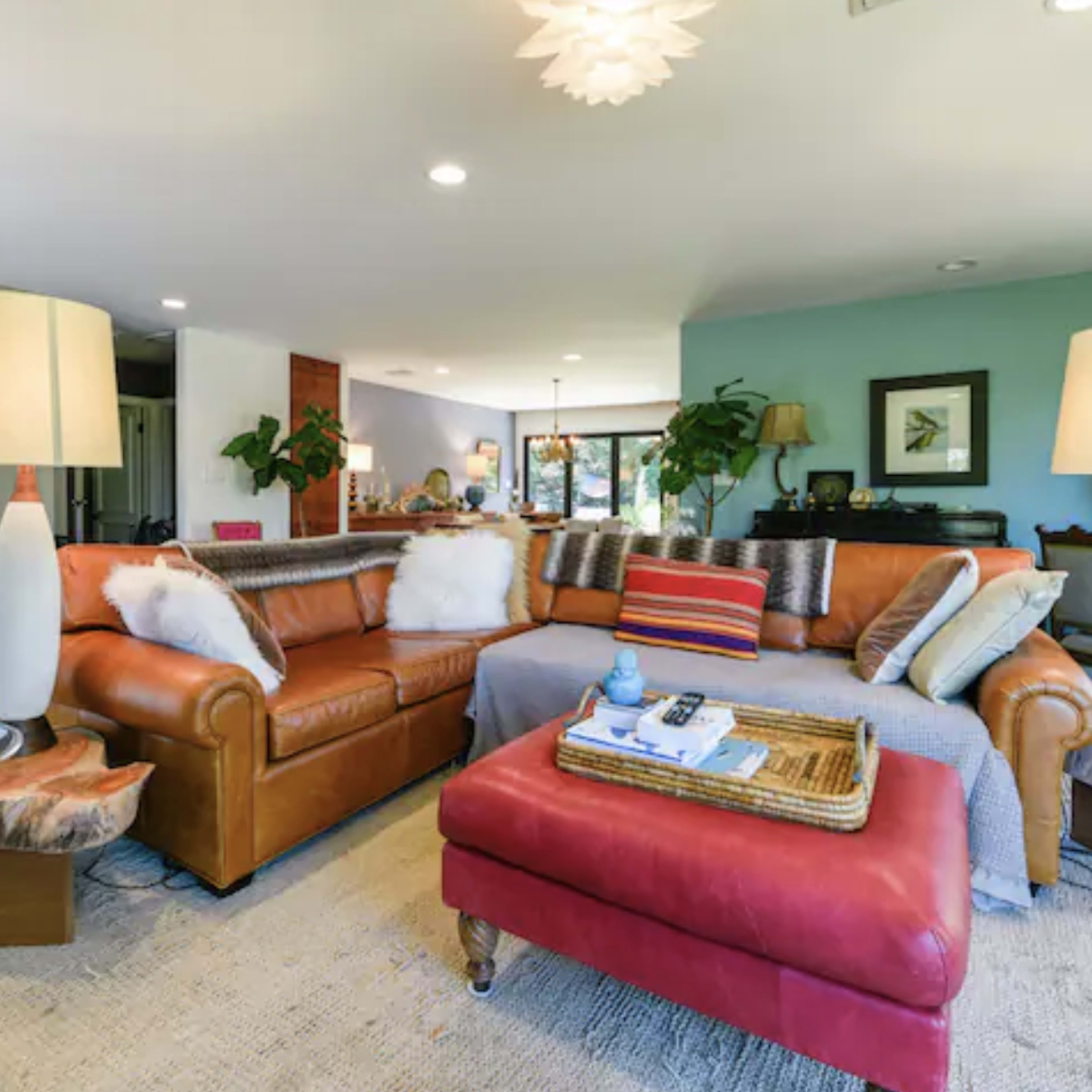 A modern living room features a brown sectional sofa with striped and patterned cushions, a red ottoman, and two tall lamps, surrounded by wooden flooring and green walls.