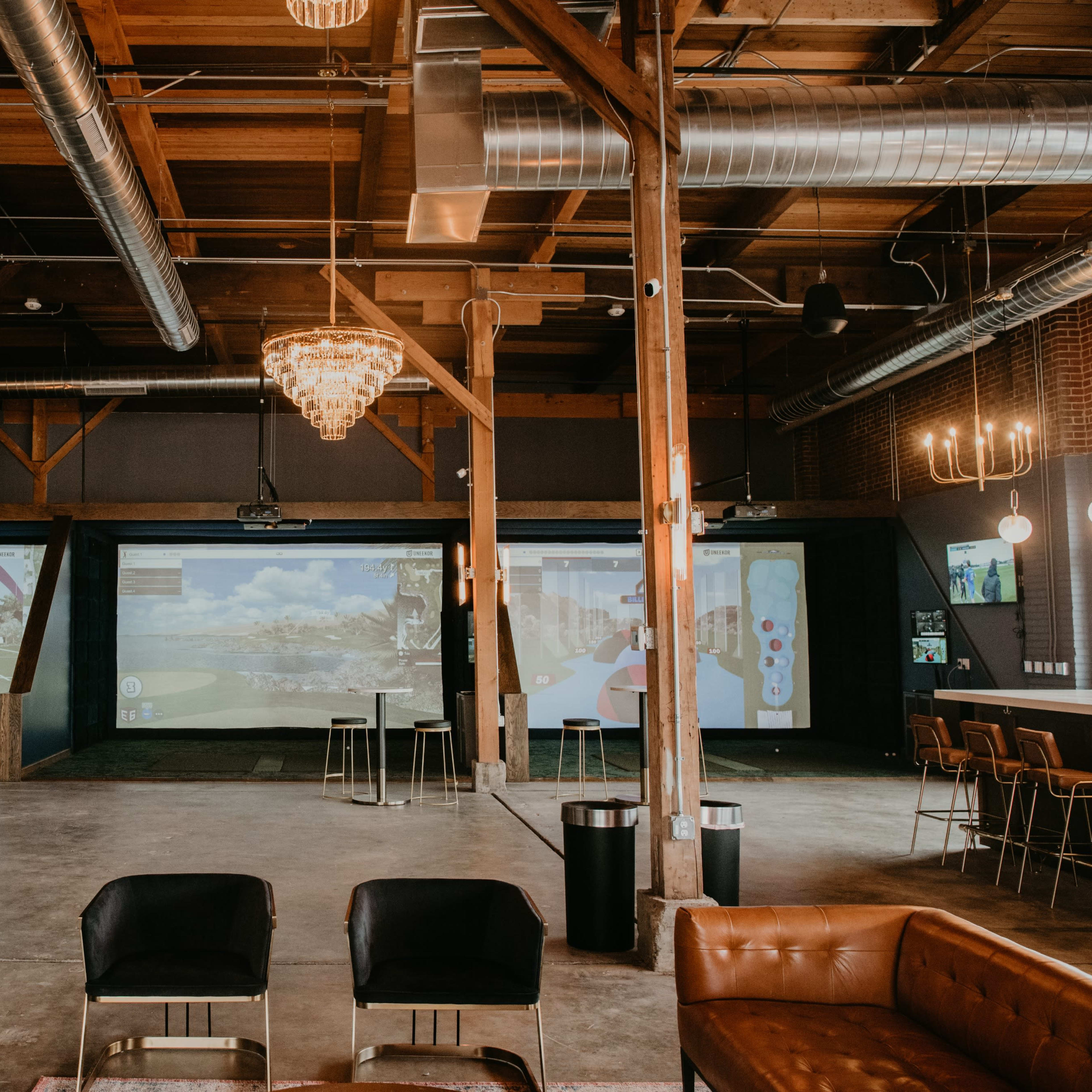 A spacious indoor venue with wooden beams, modern lighting, and multiple large screens displaying virtual golf simulations.