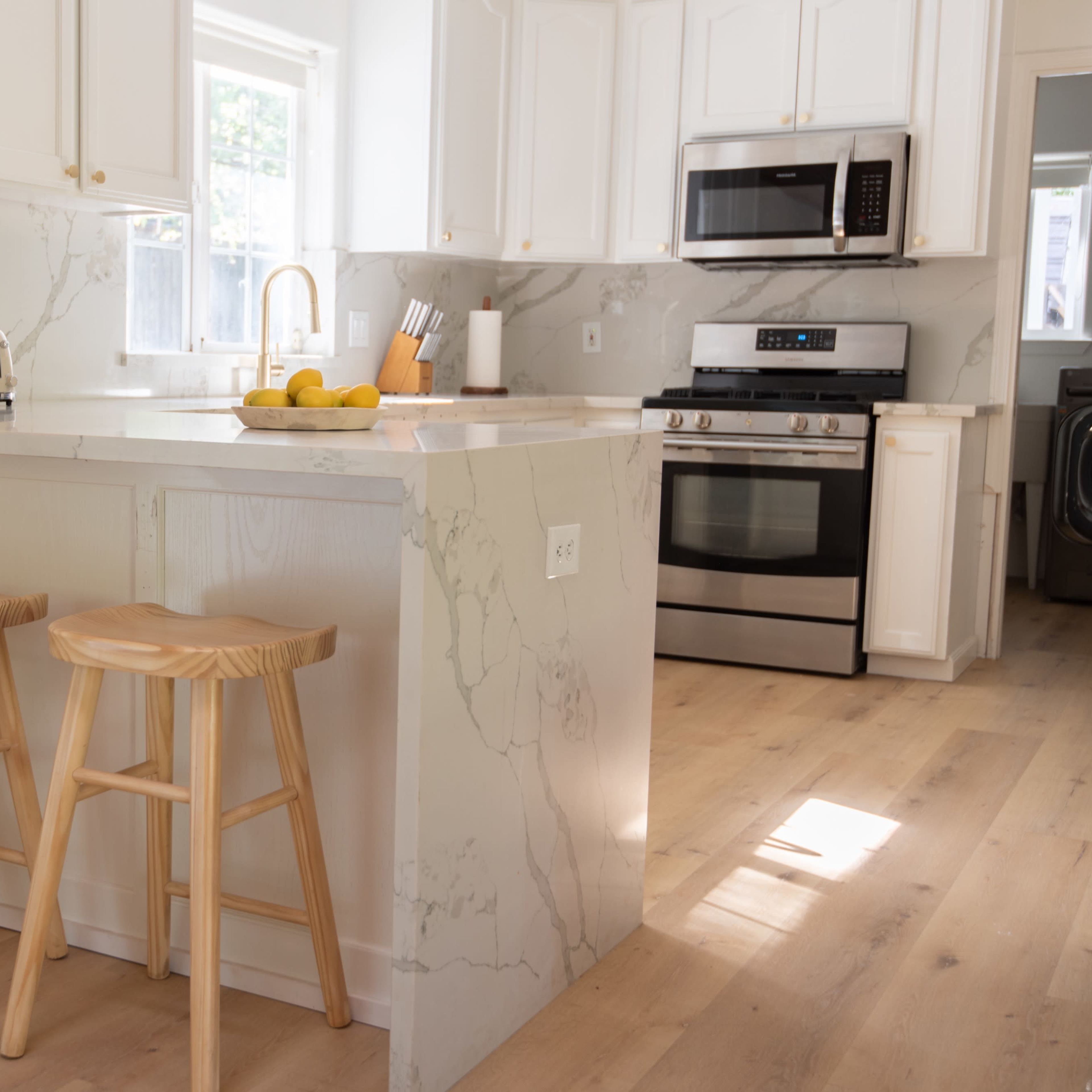 A modern kitchen features white cabinetry, a marble countertop island with three wooden stools, and stainless steel appliances.