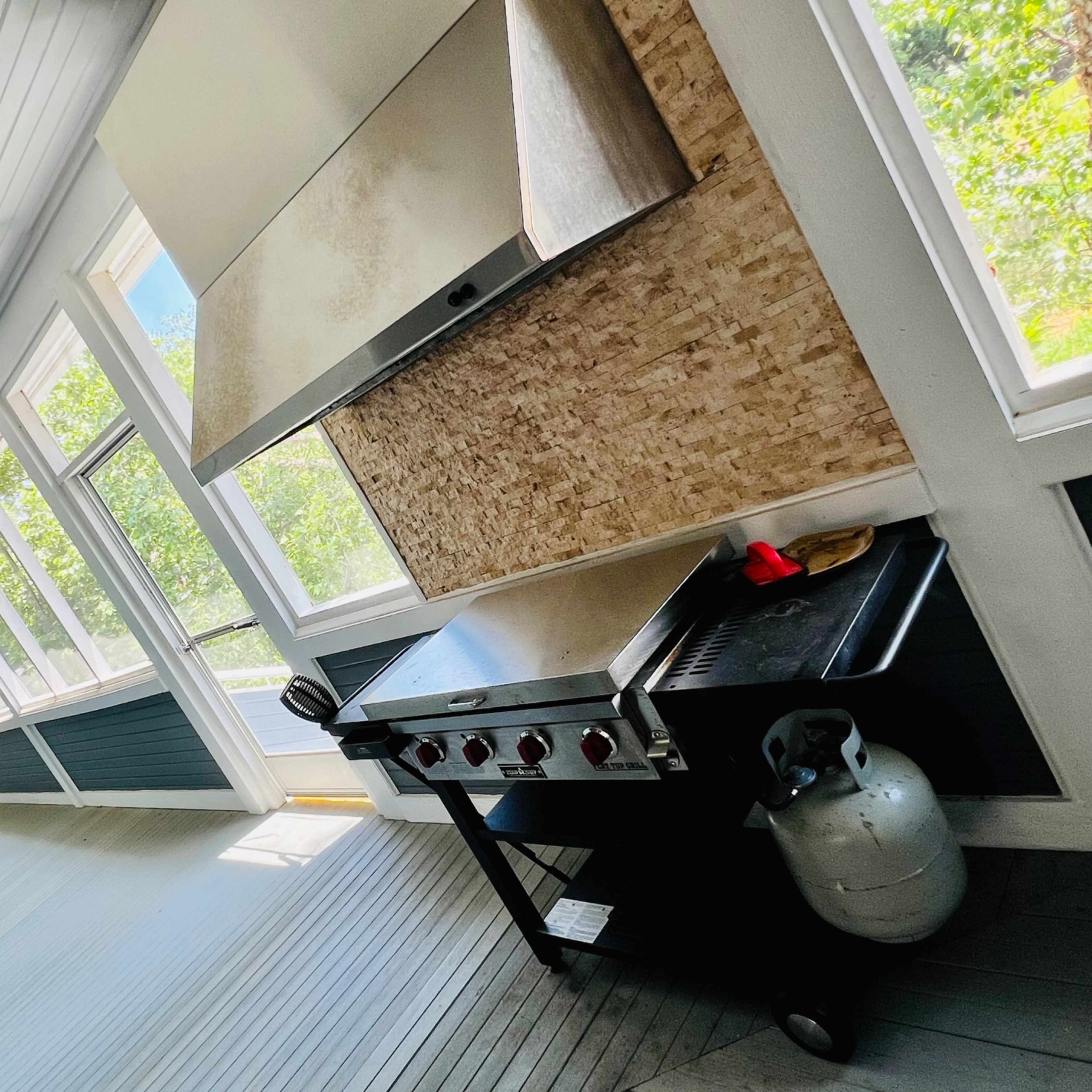 A spacious, sunlit porch featuring a stainless steel grill and a propane tank, with large windows allowing natural light to enter.