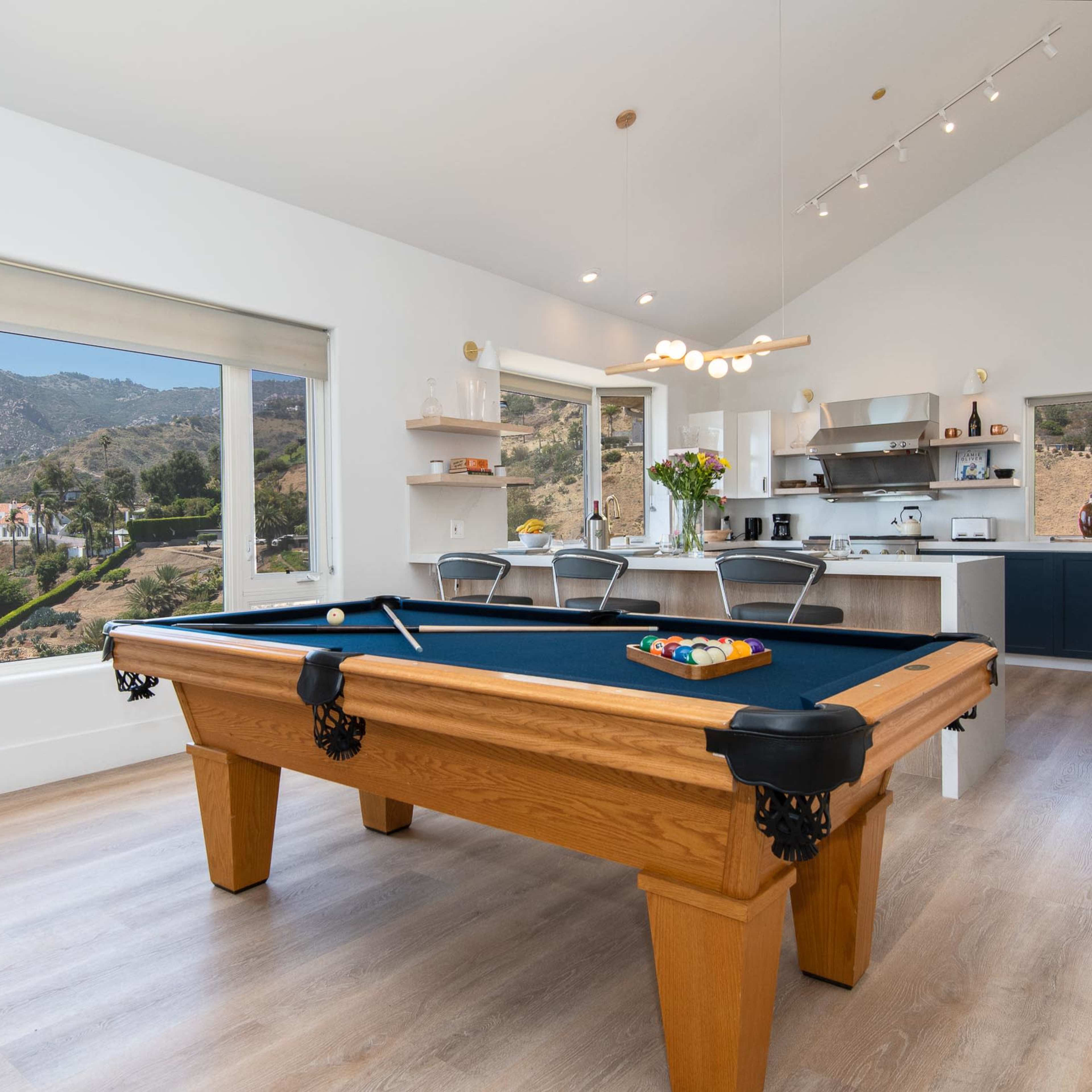A modern game room featuring a pool table, large windows with a mountain view, and an open kitchen area.