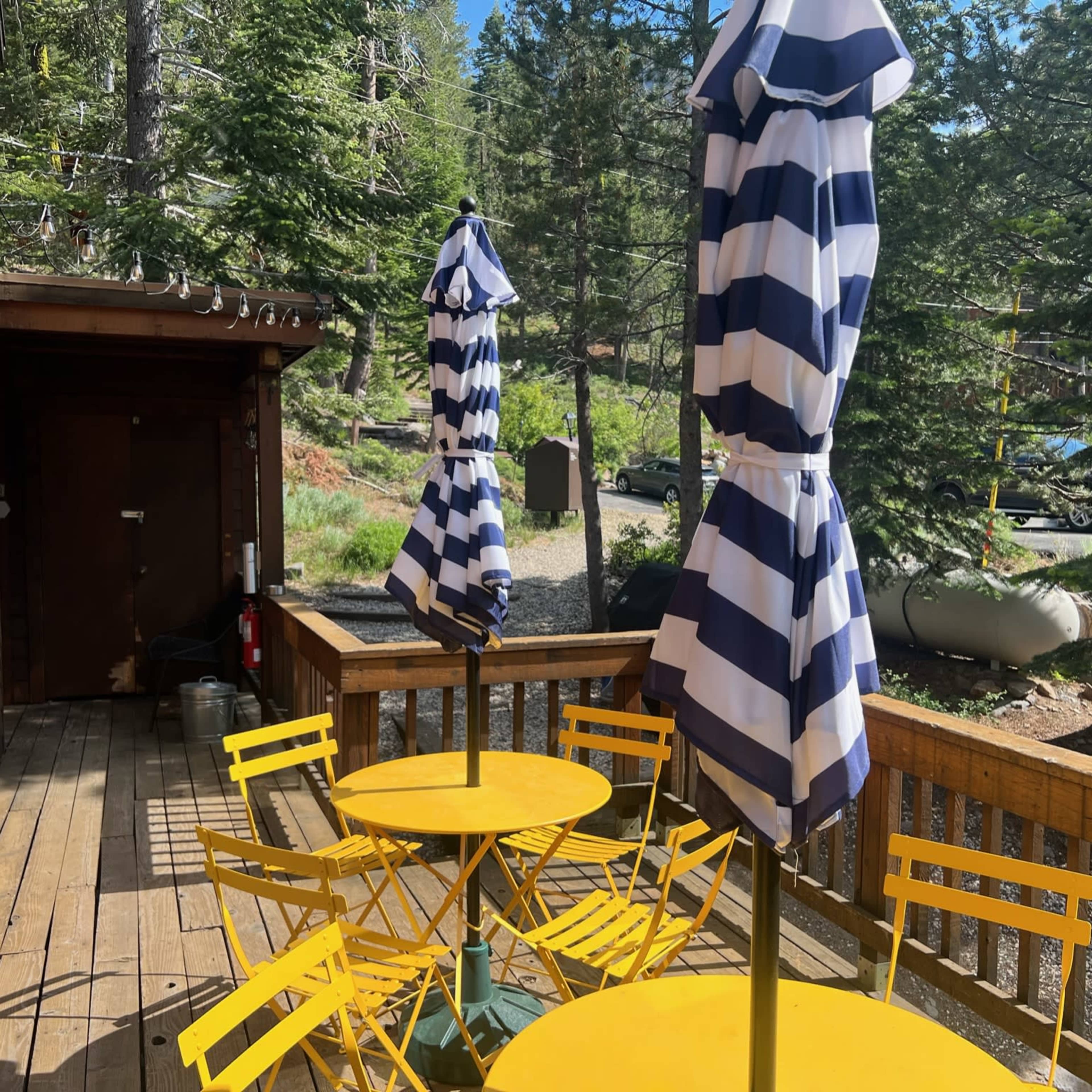 A wooden deck features yellow tables and blue-and-white striped umbrellas surrounded by trees.