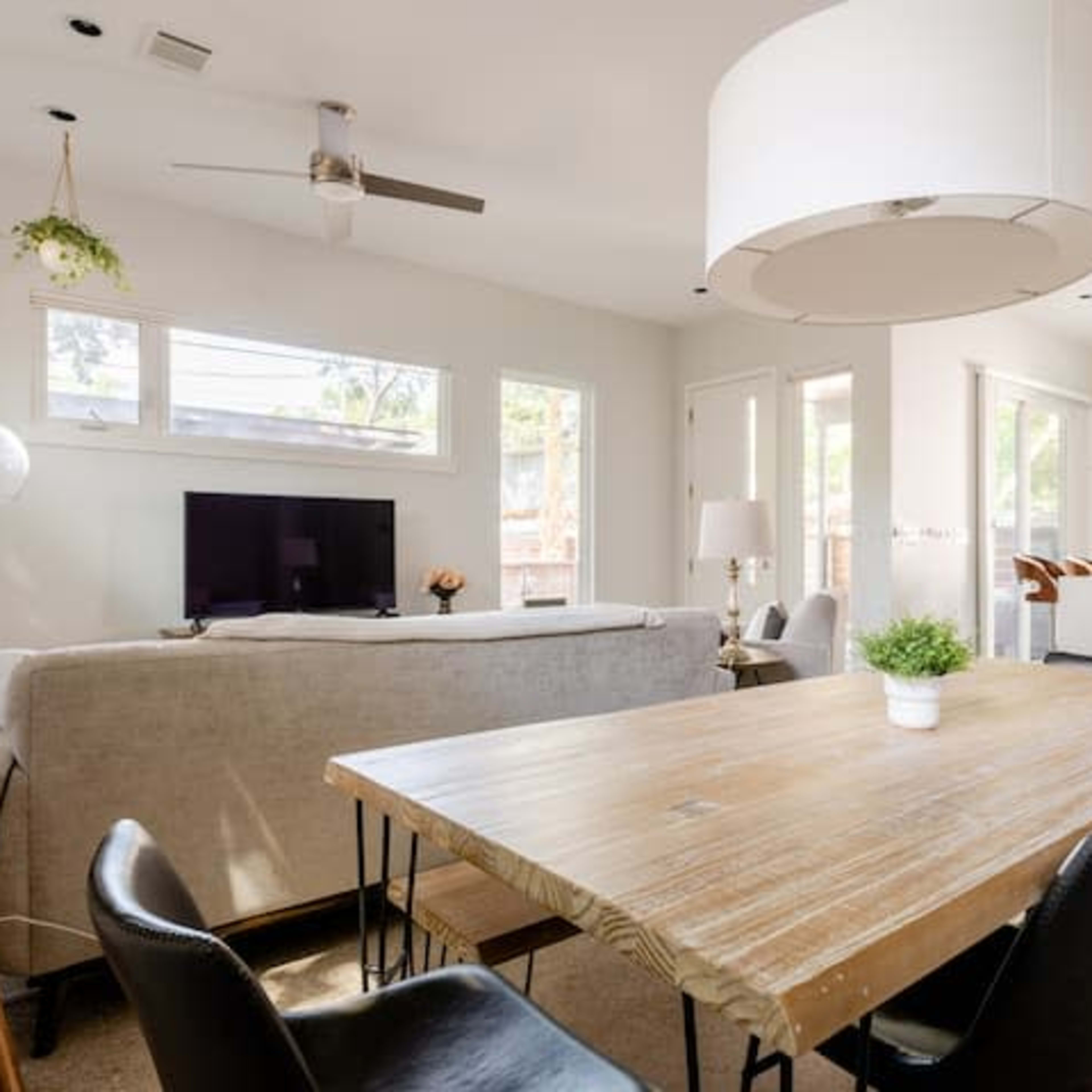 This image shows a modern living and dining area with a wooden dining table, black chairs, and a light-colored sofa facing a television.