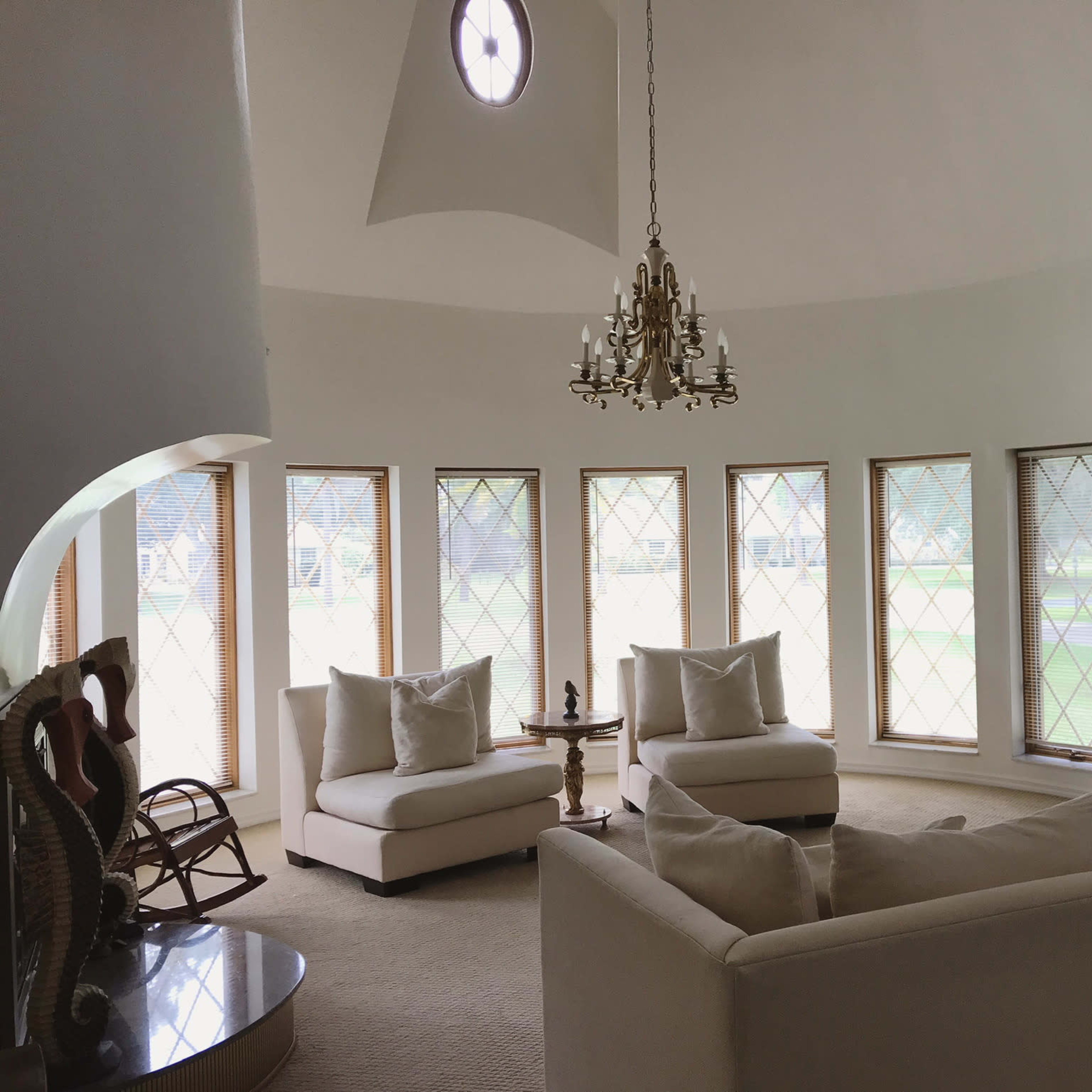 A circular living room features a chandelier, two armchairs, and a large sectional couch arranged around a coffee table, with tall windows providing natural light.