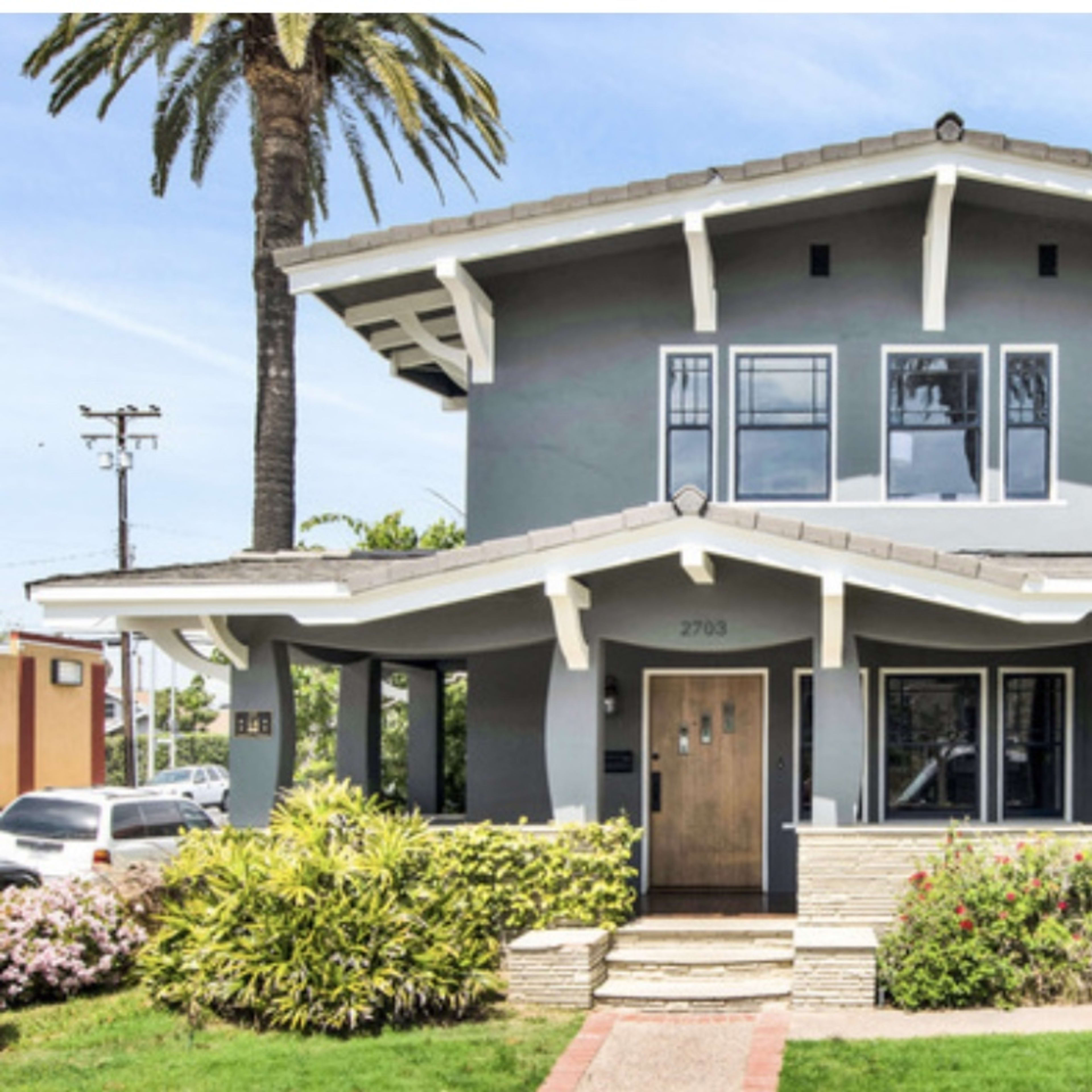 A two-story gray house features a wide front porch, multiple windows, and landscaped front gardens with palm trees nearby.