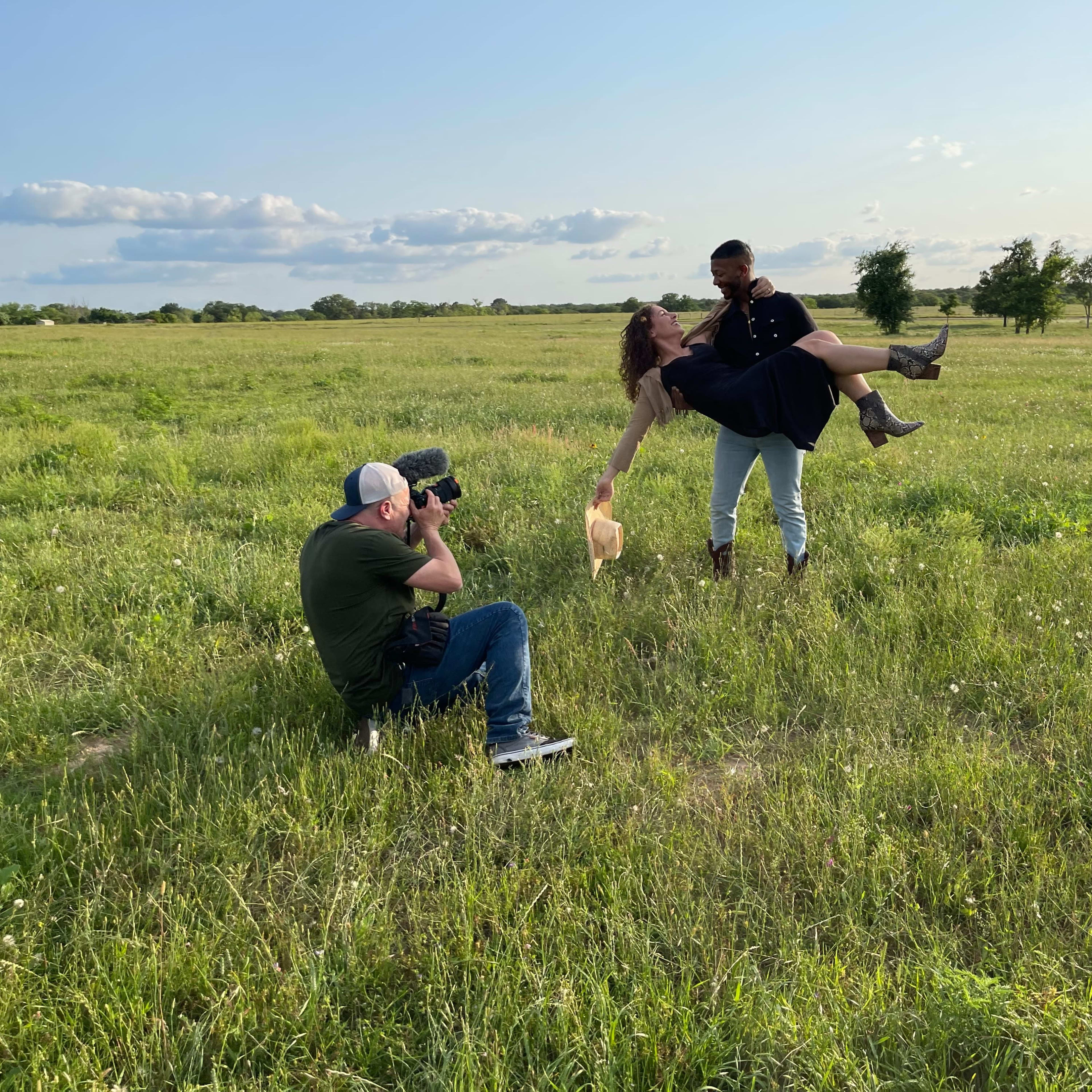 A photographer captures a couple in a field, with one partner holding the other in a playful pose.