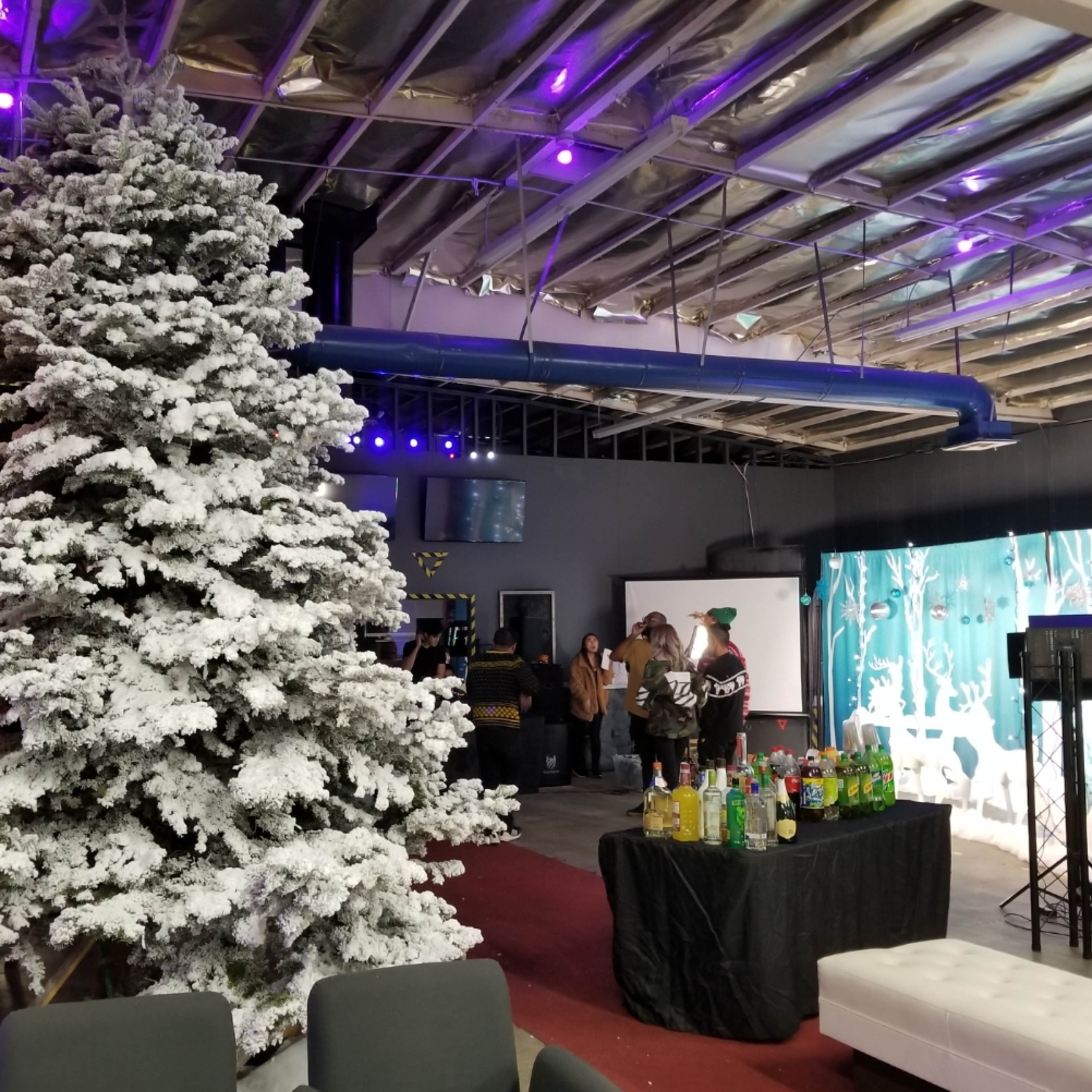A large, artificial snow-covered Christmas tree stands next to a bar table filled with colorful drinks in a festive indoor space.