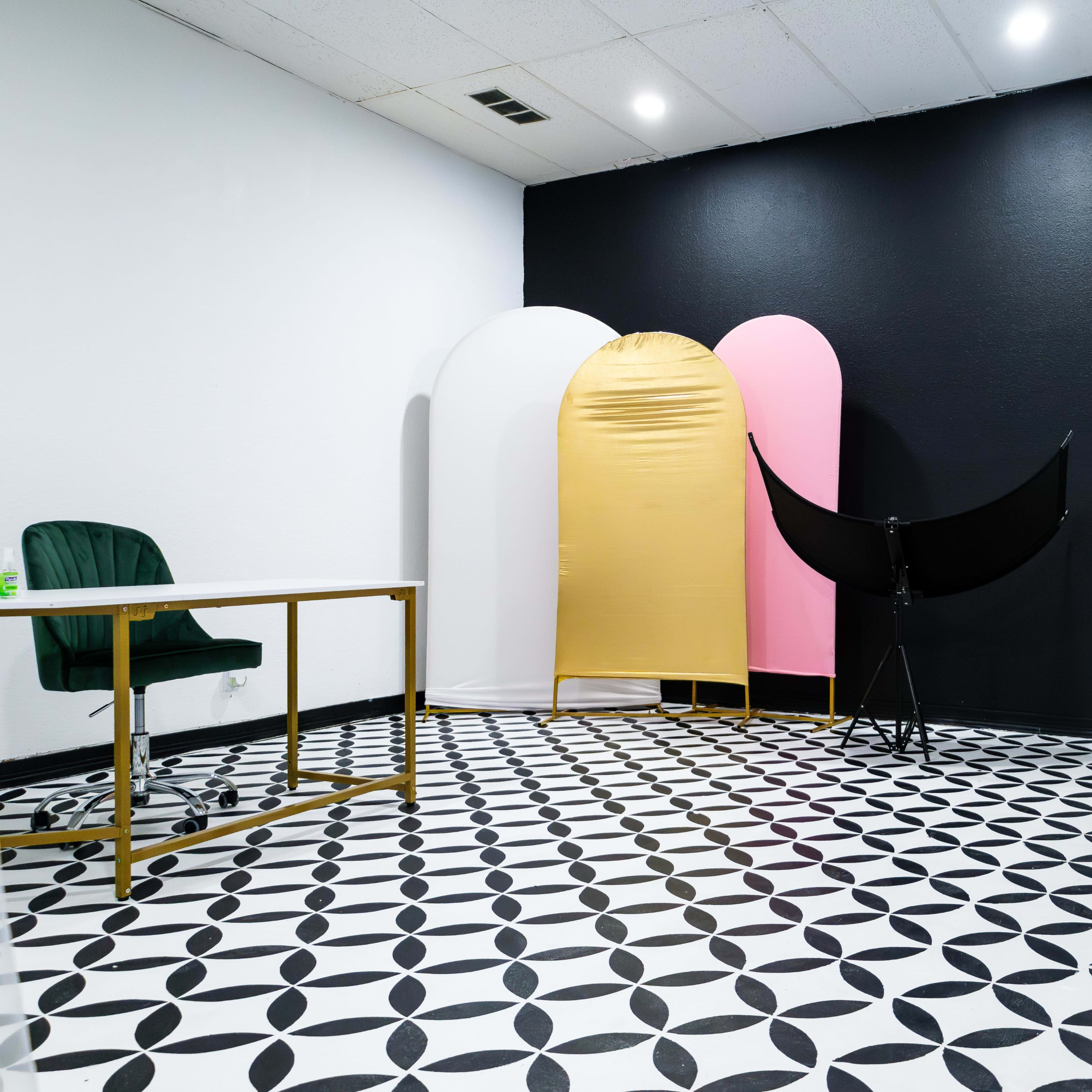 A modern studio space with patterned black and white tile flooring, a gold, pink, and white backdrop setup, and a folding screen, along with a green chair and a table.