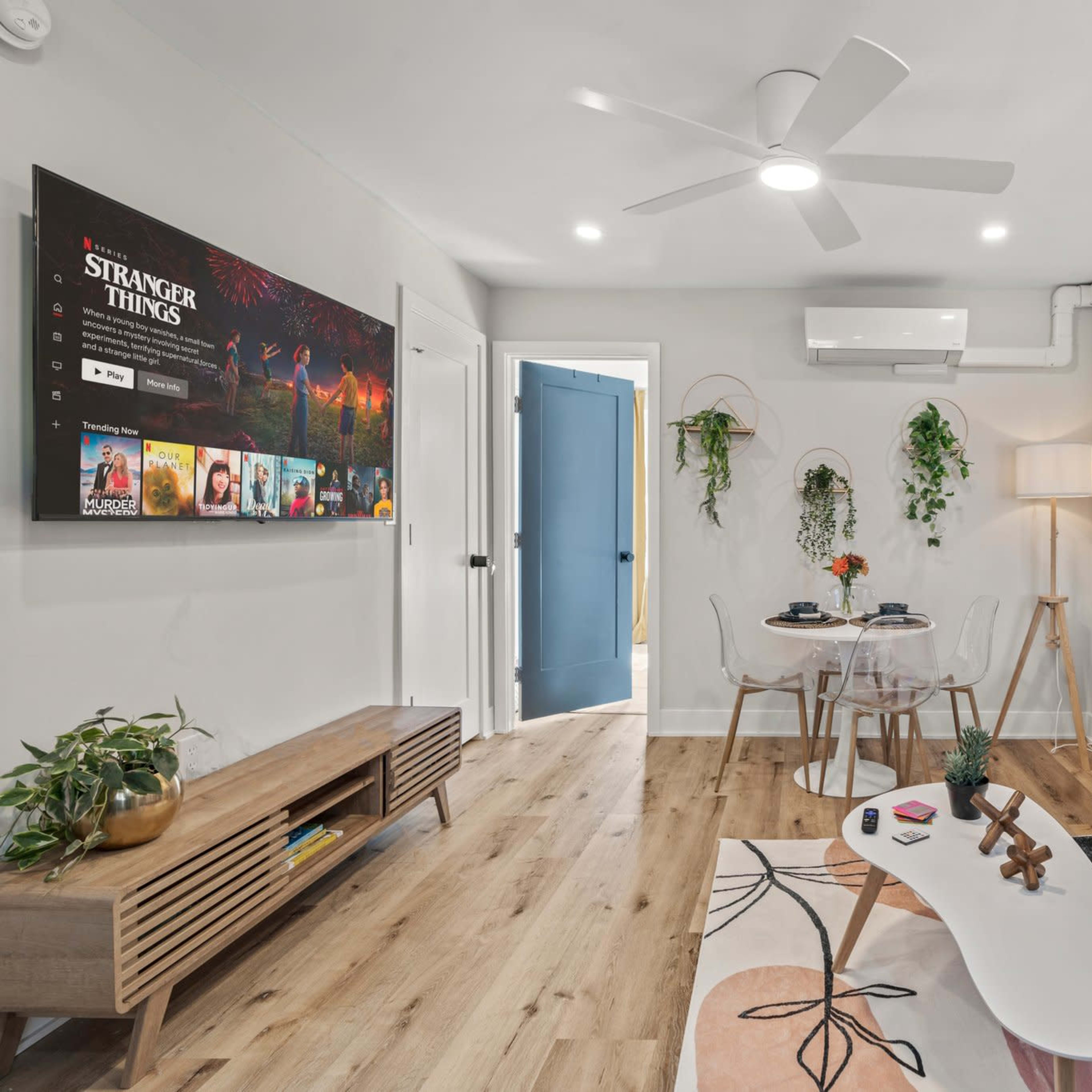 A modern living room with a light gray sofa, a coffee table, a wooden TV unit displaying a "Stranger Things" poster, and decor featuring plants against a white wall.