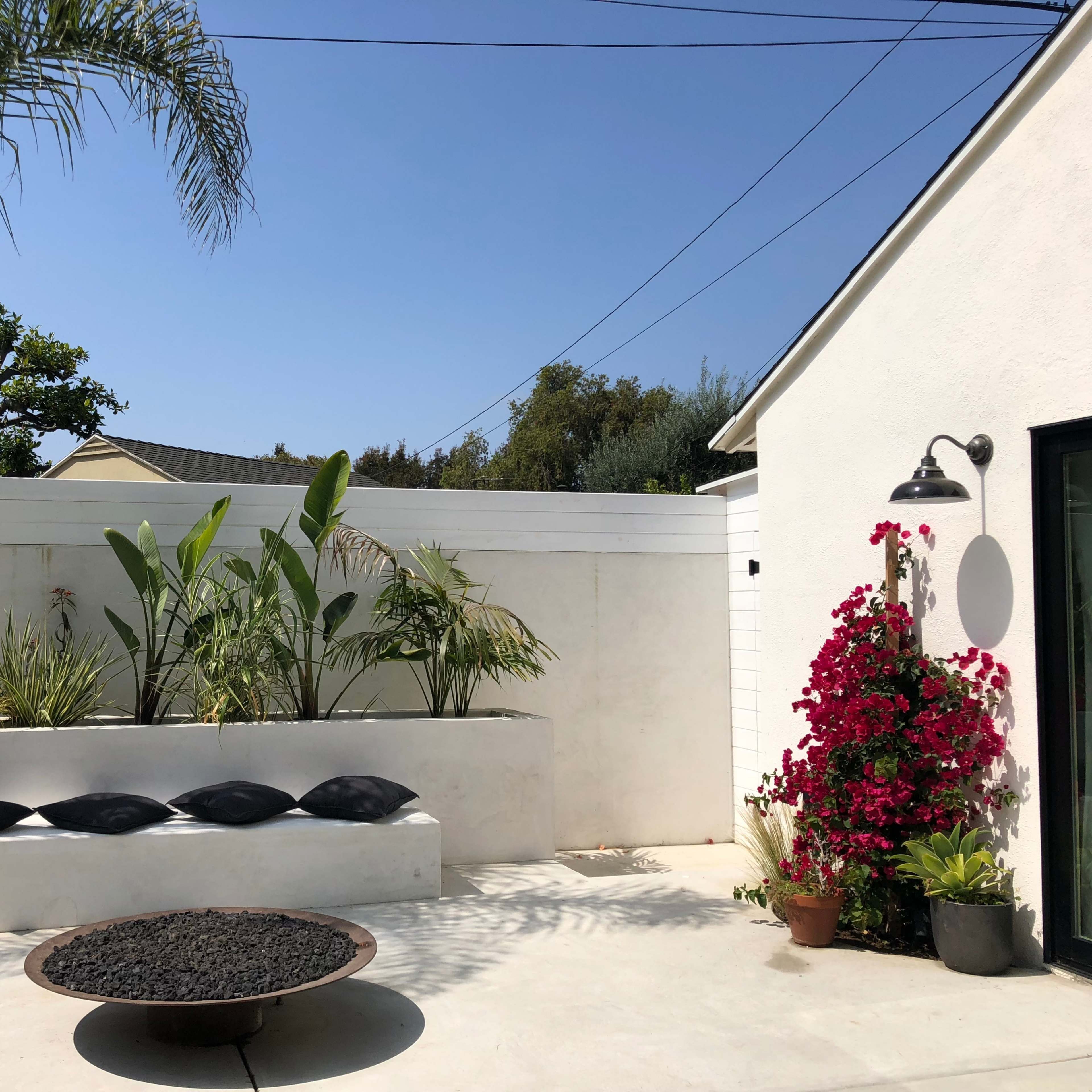 A modern outdoor space featuring a concrete planter with greenery, a round fire pit, and a wall adorned with vibrant flowers against a clear blue sky.