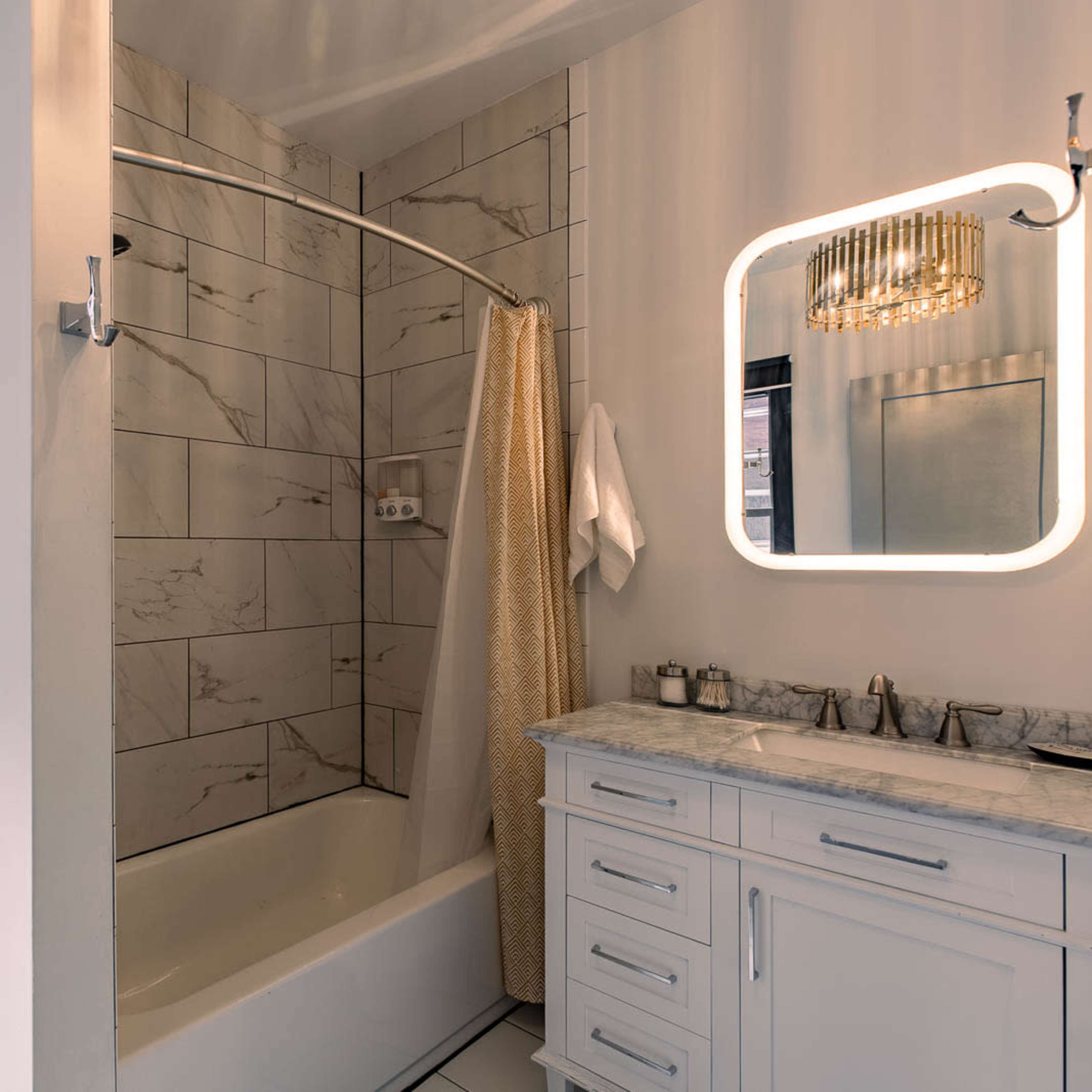 A bathroom featuring a bathtub with a shower curtain, a toilet, a vanity with a marble countertop, and a round mirror illuminated by a light fixture.