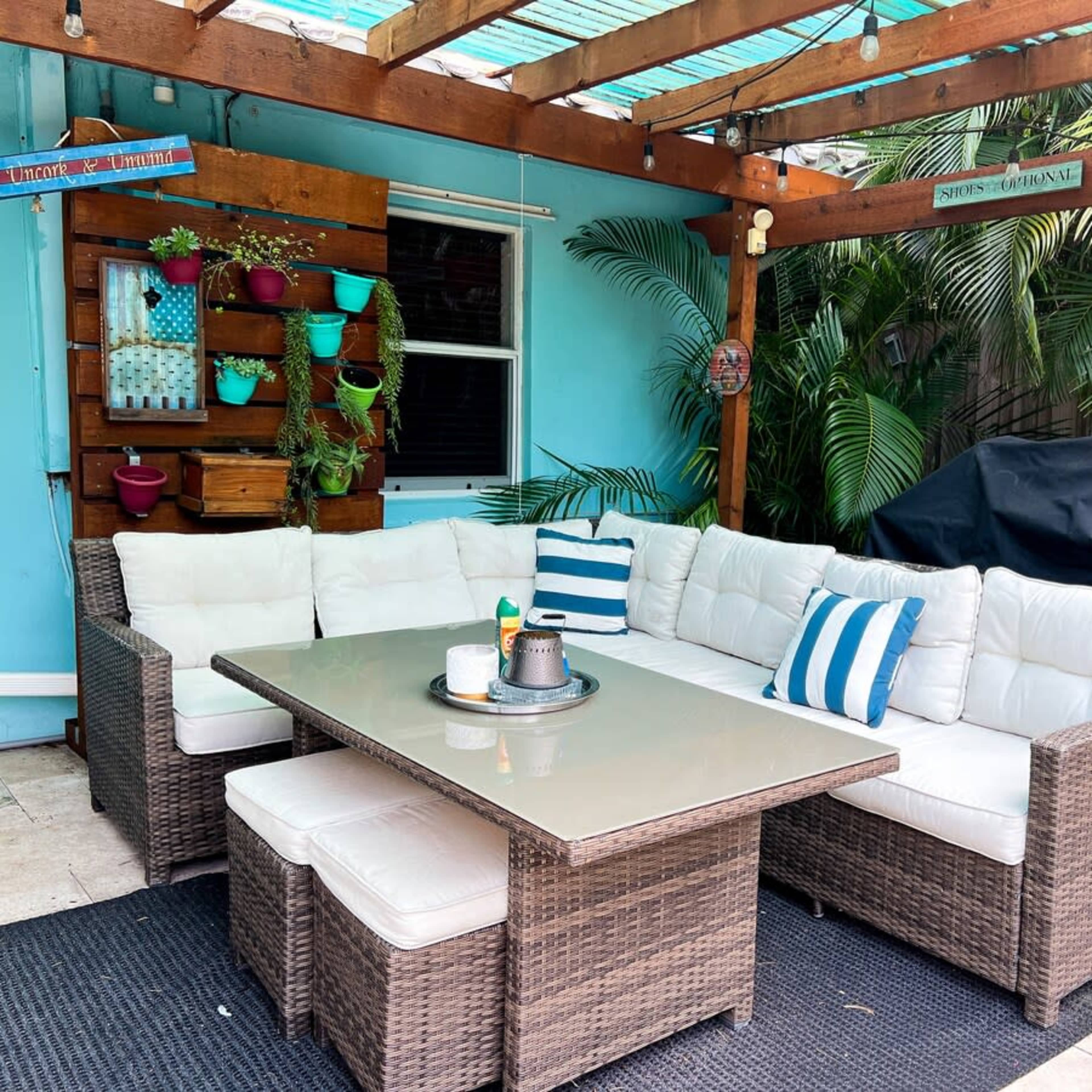 A covered outdoor seating area features a large sectional sofa, a glass-topped table, and decorative potted plants against a blue wall.
