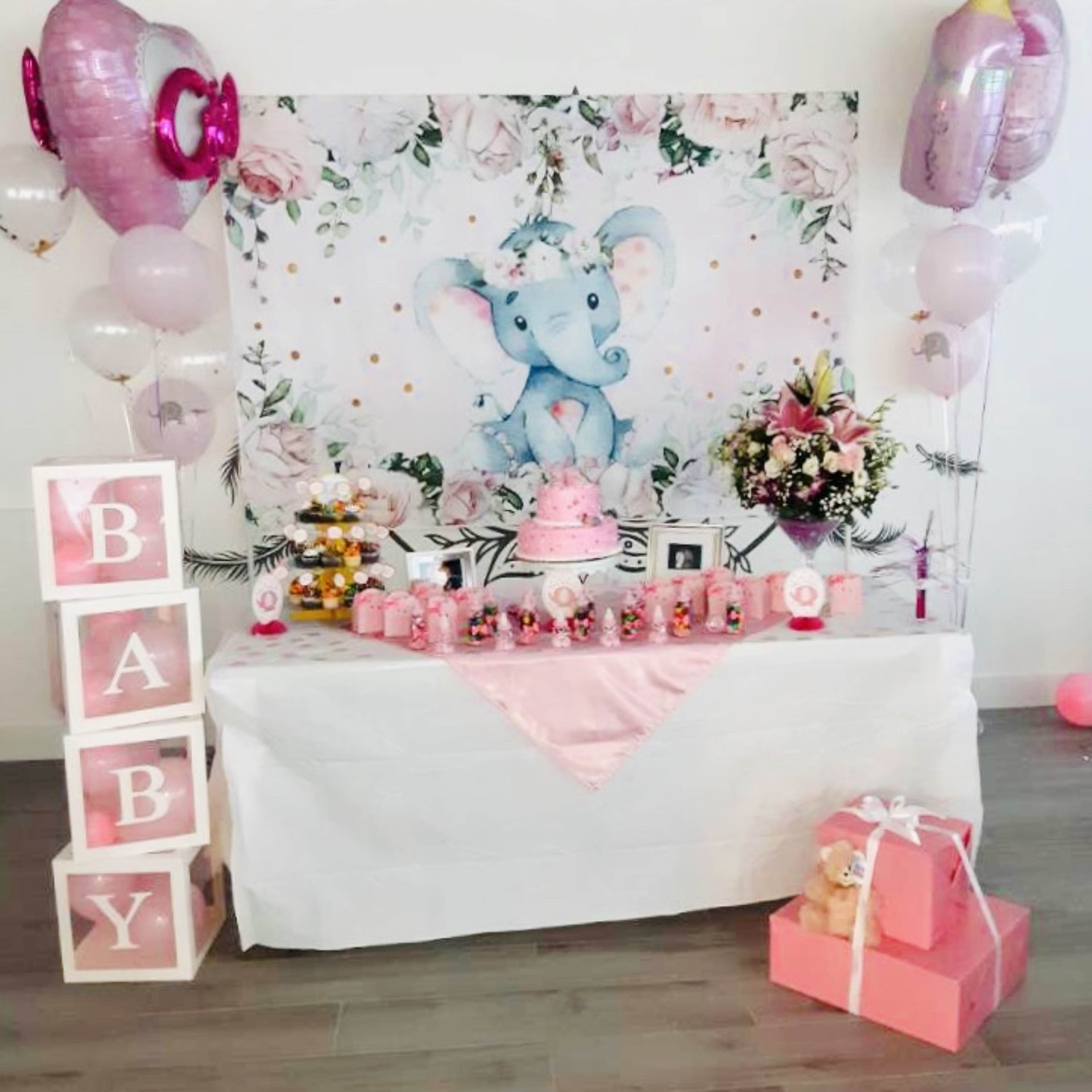 A decorated table features a pink-themed baby shower setup with an elephant backdrop, a cake, gifts, and floral arrangements.