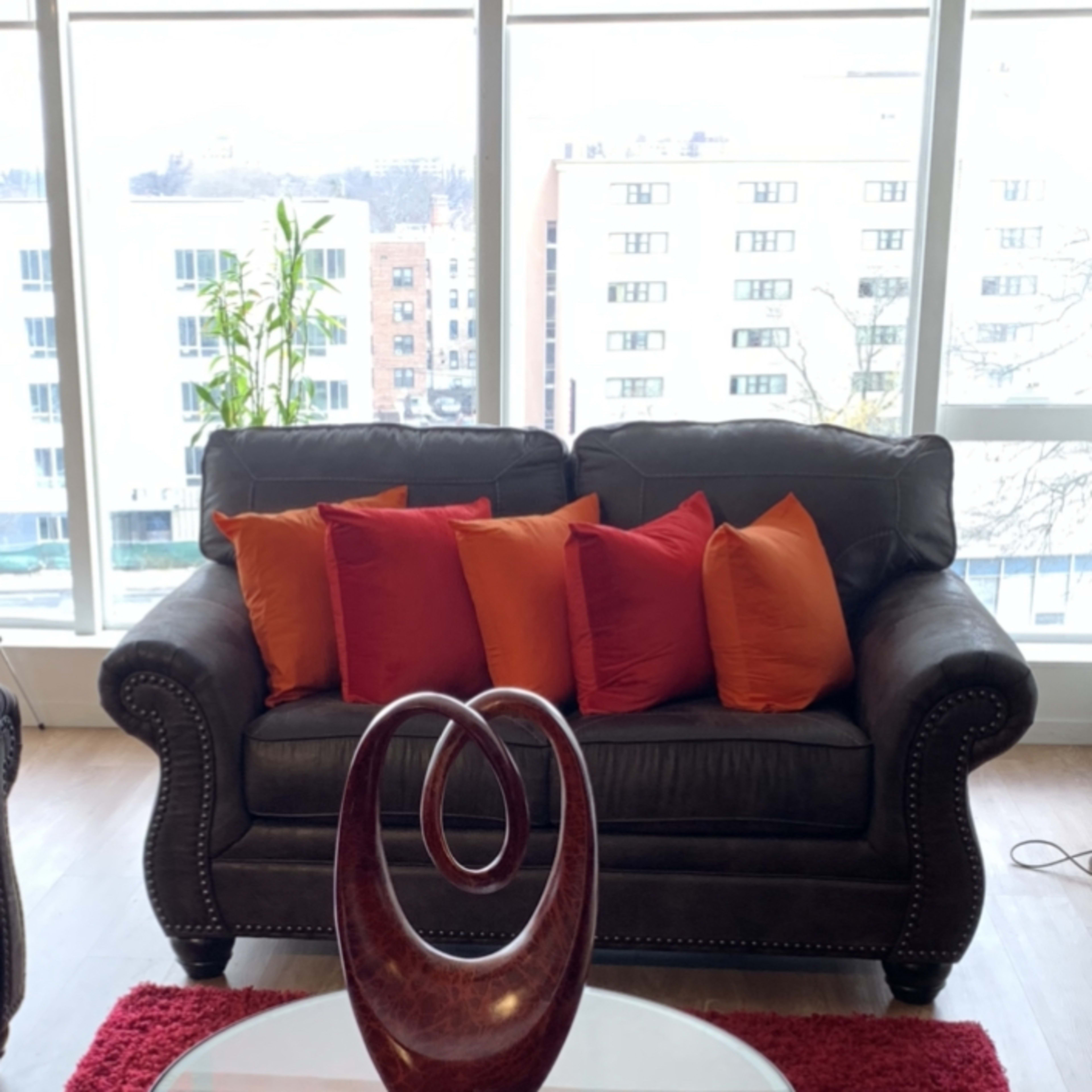 A leather couch with orange and red pillows sits in front of a glass coffee table on a red rug, with a decorative wooden sculpture and a candle on the table.