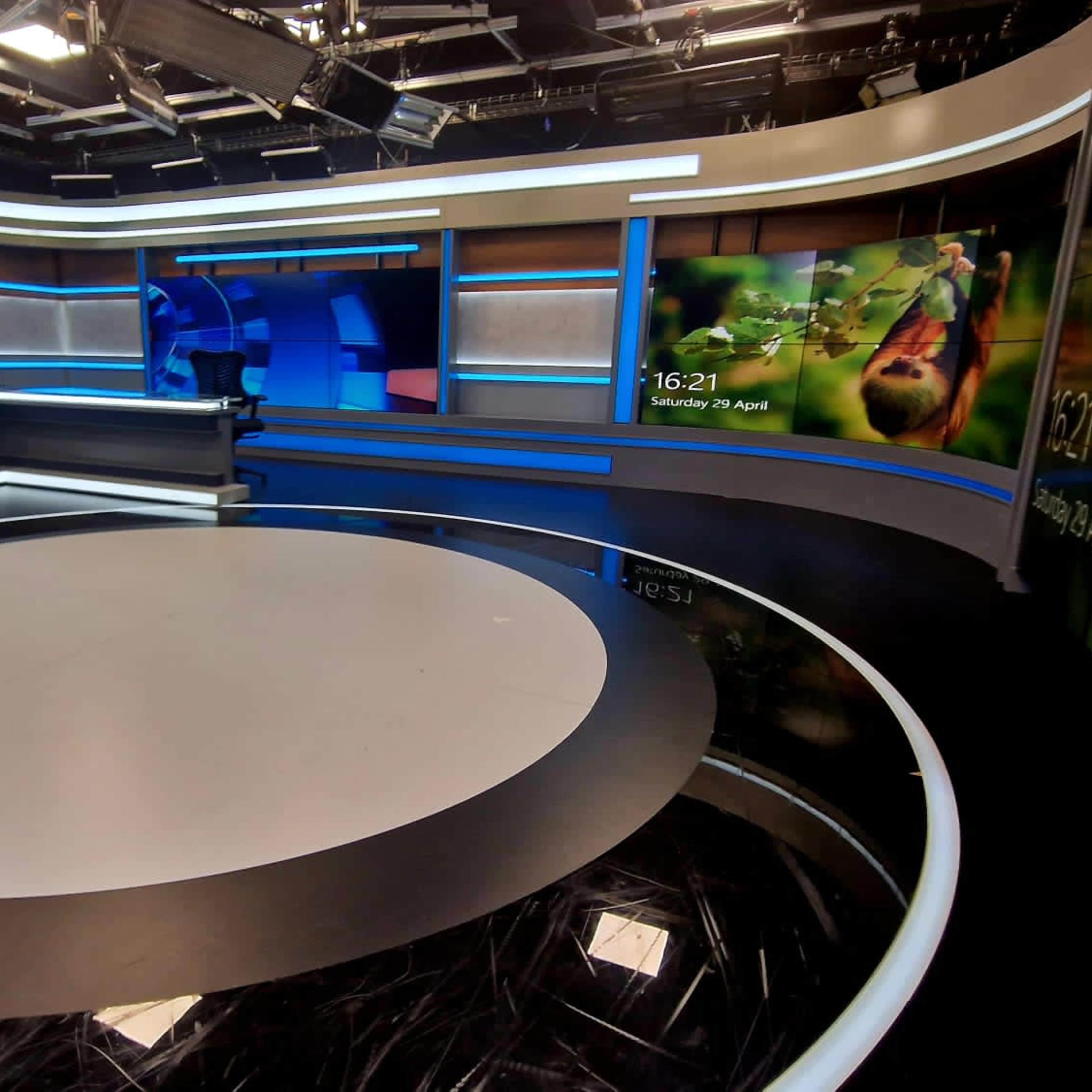 A modern news studio with a circular stage area, large screens displaying information, and a backdrop featuring a nature scene.