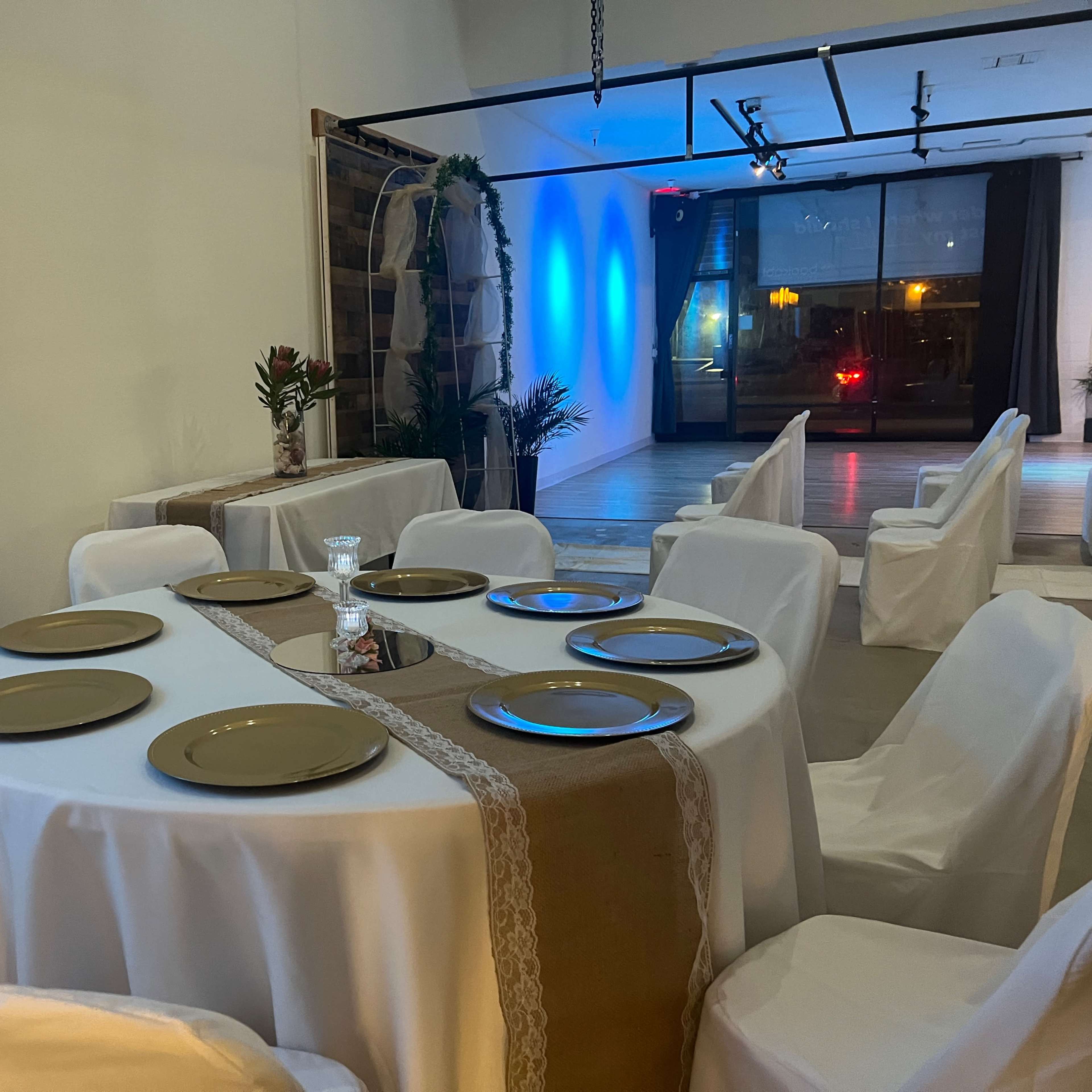 A round table with eight gold plates is set for a meal in a dimly lit dining area that features white draped chairs and blue lighting.