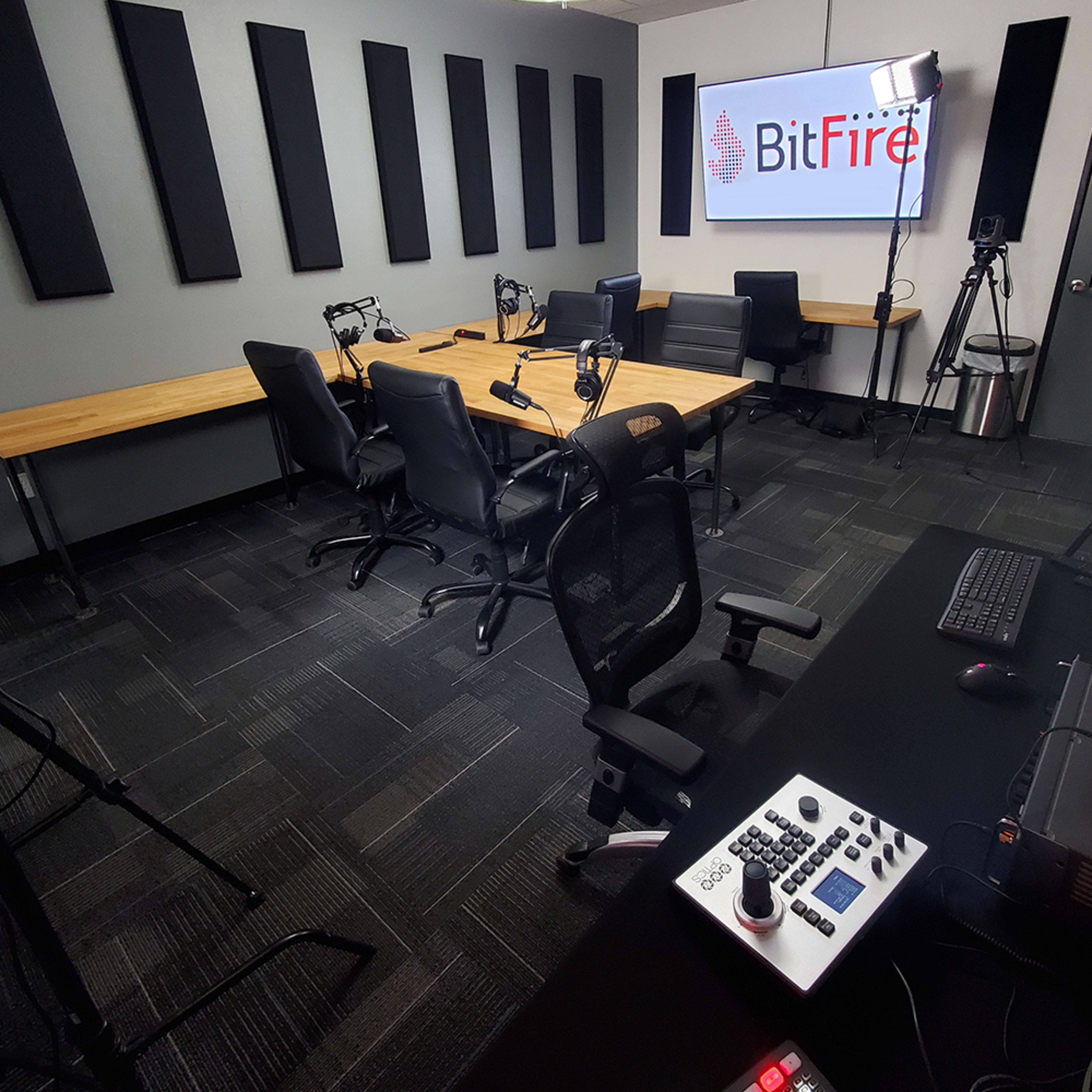 A modern podcast studio with a large table, multiple chairs, microphones, and audio equipment, featuring a screen displaying the BitFire logo.