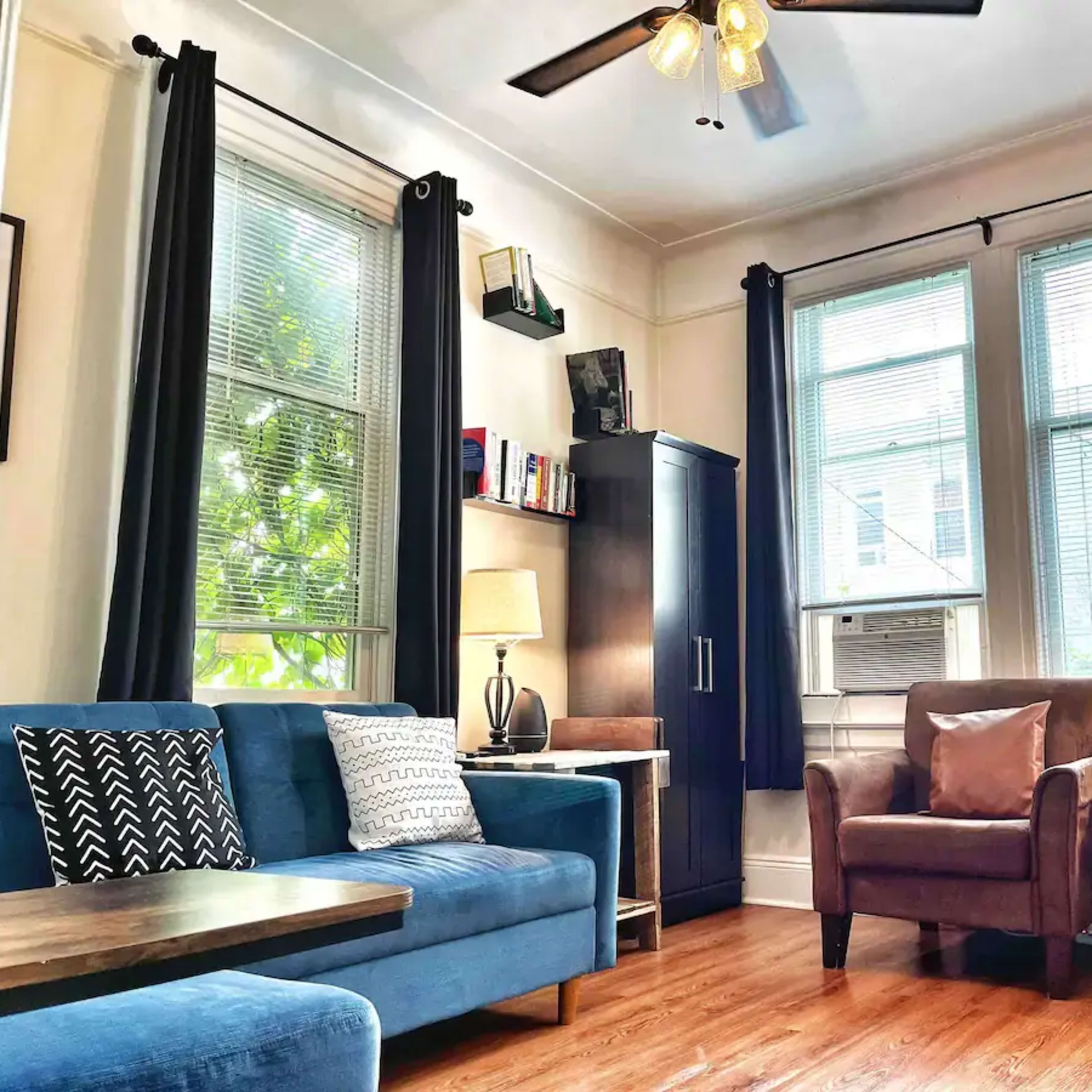A cozy living room featuring blue and brown furniture, large windows with natural light, and a mixture of lamps and shelves for décor.