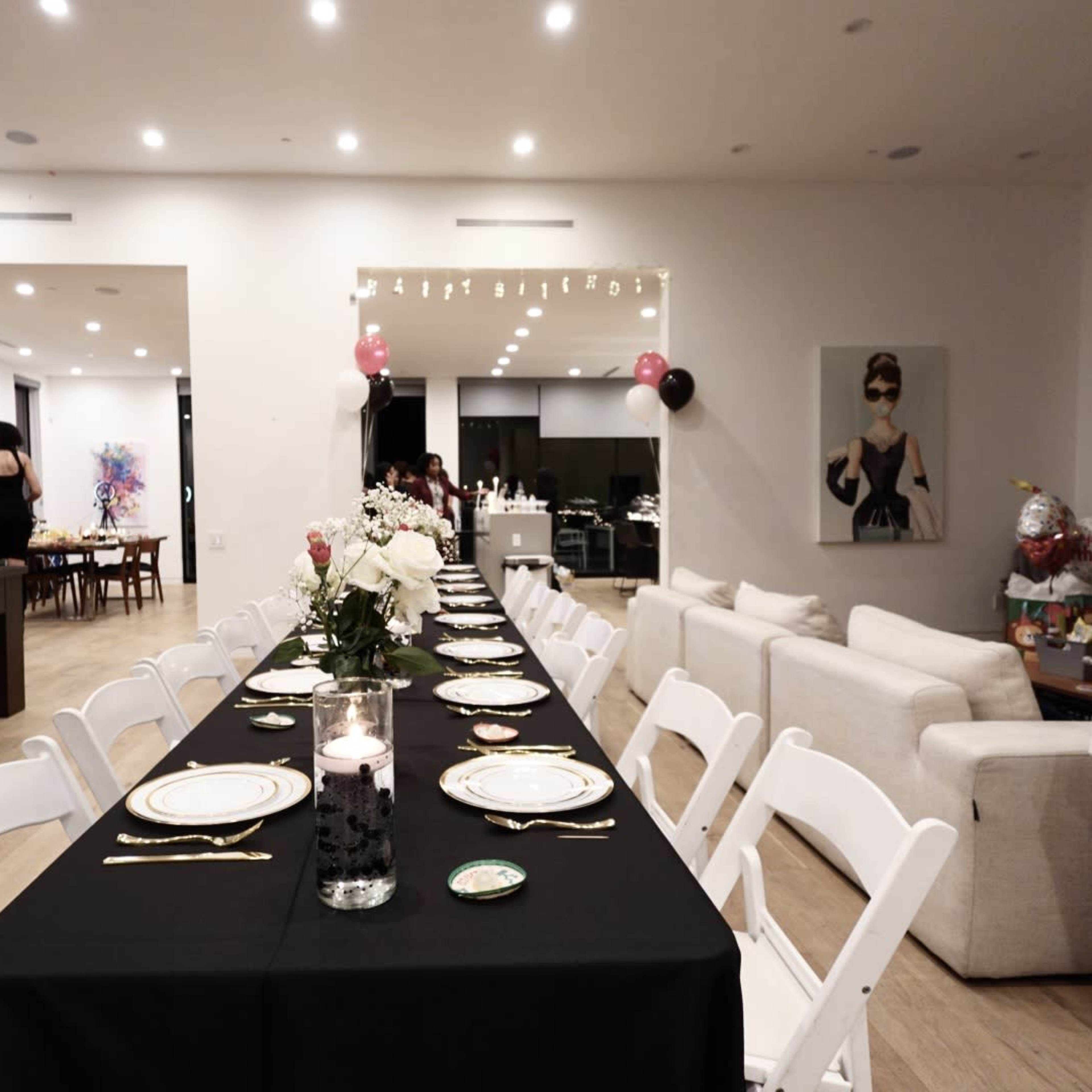 A long black table is set for a dinner party in a modern dining area, with white chairs and decor including flowers and candles, alongside a pool table and an open kitchen.