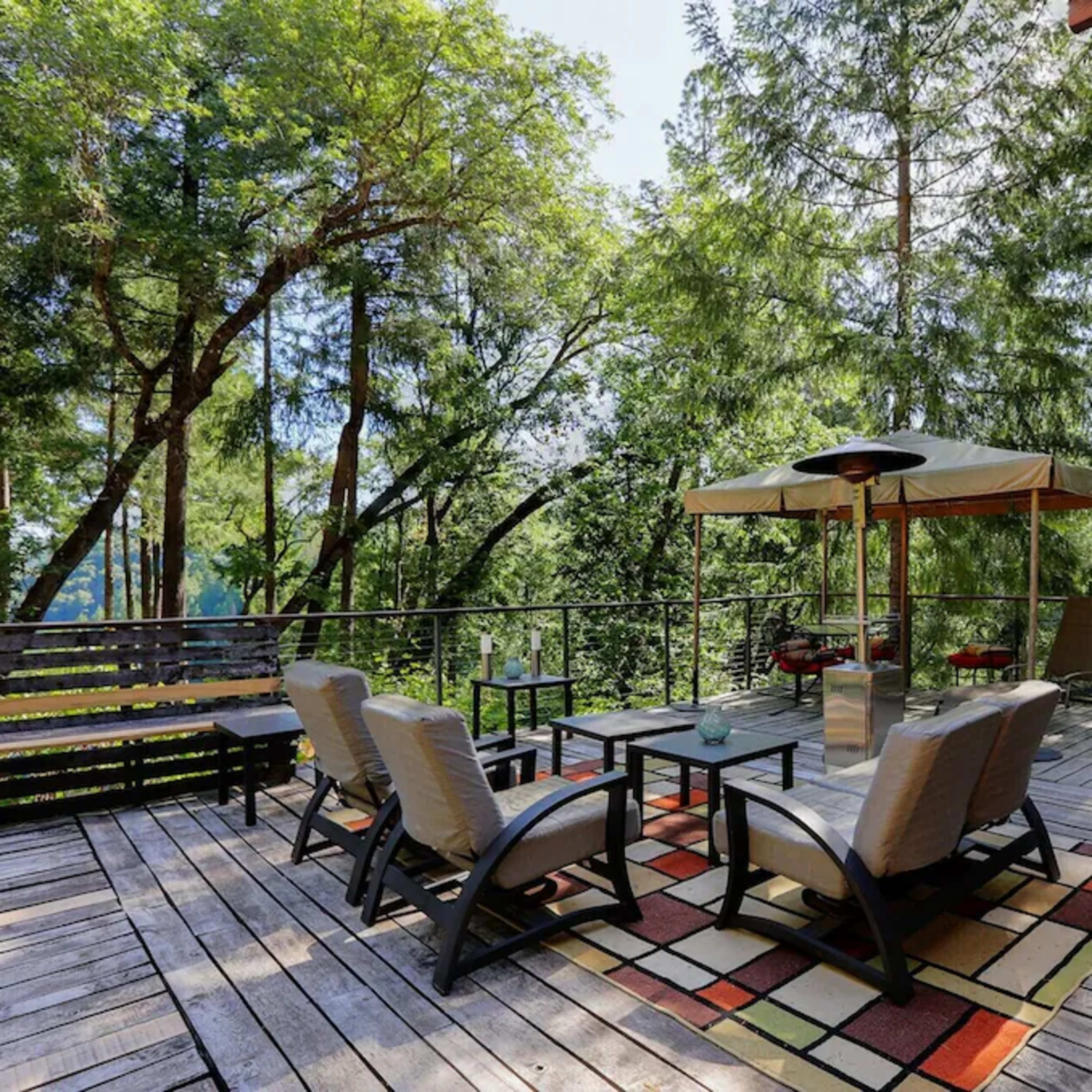 A wooden deck surrounded by trees, featuring seating arrangements, a canopy, and a grill.