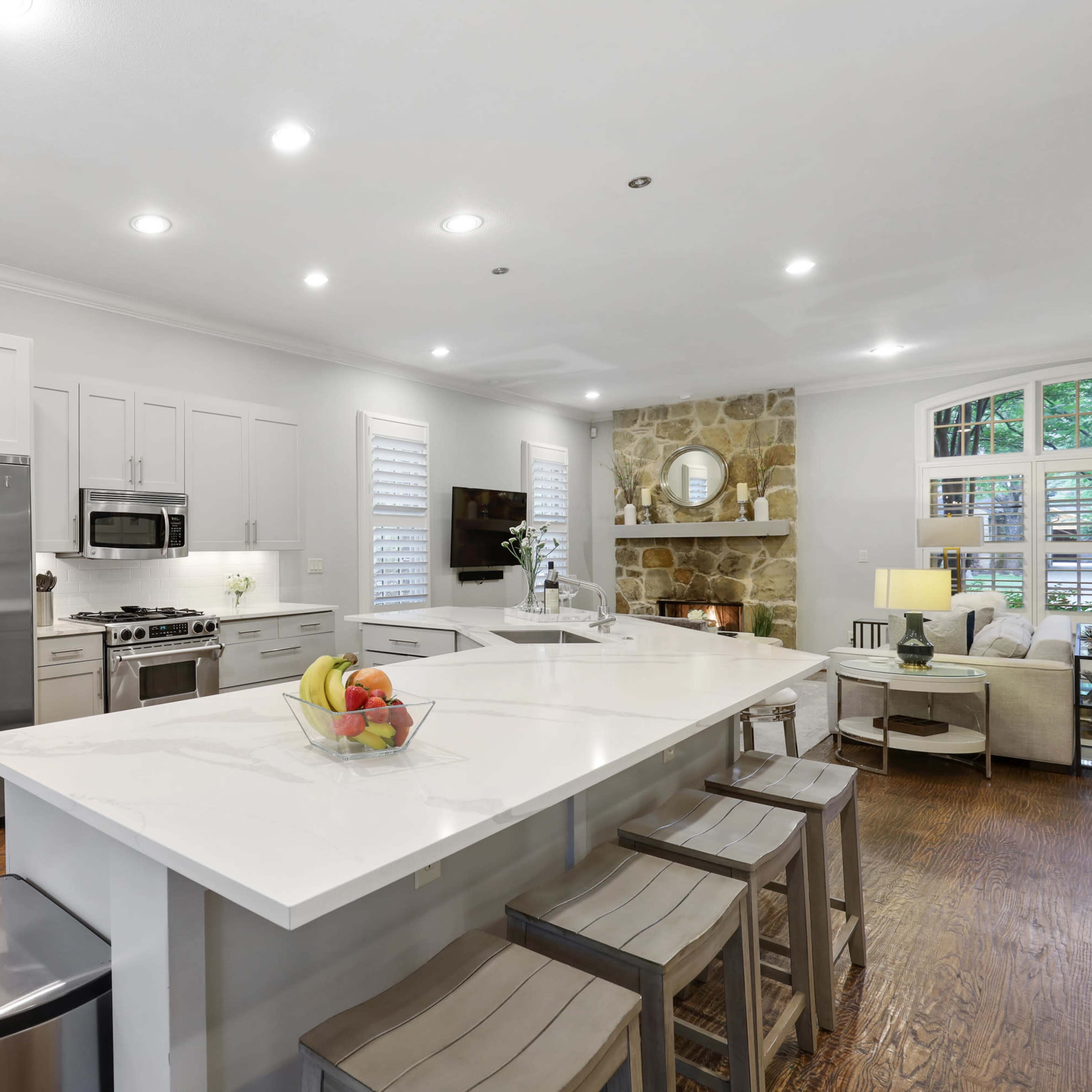 A modern kitchen with gray cabinetry, stainless steel appliances, a white island with bar stools, and a cozy seating area near a stone accent wall.