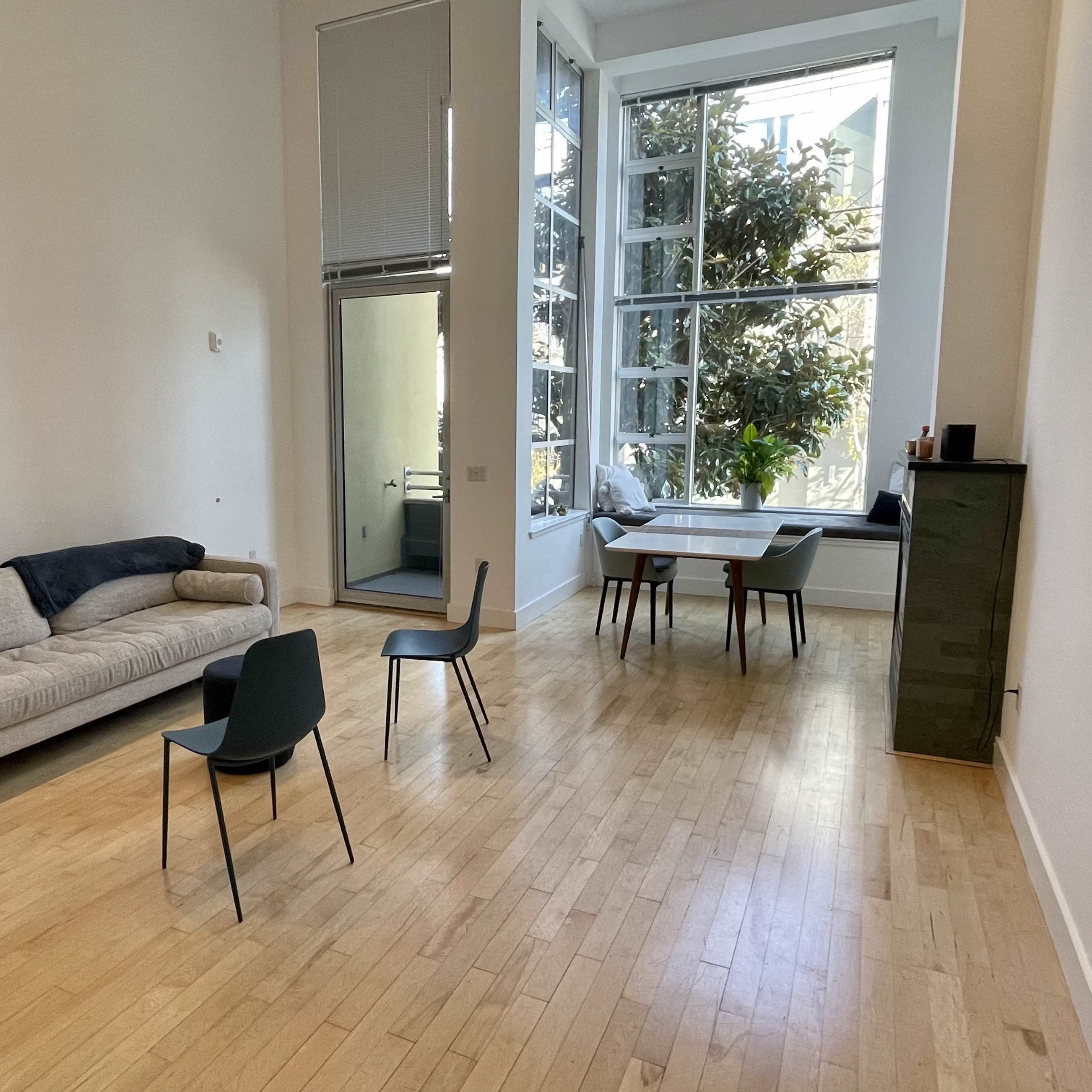 A bright, minimalist room with a sofa, a table with chairs, and large windows allowing natural light to enter.